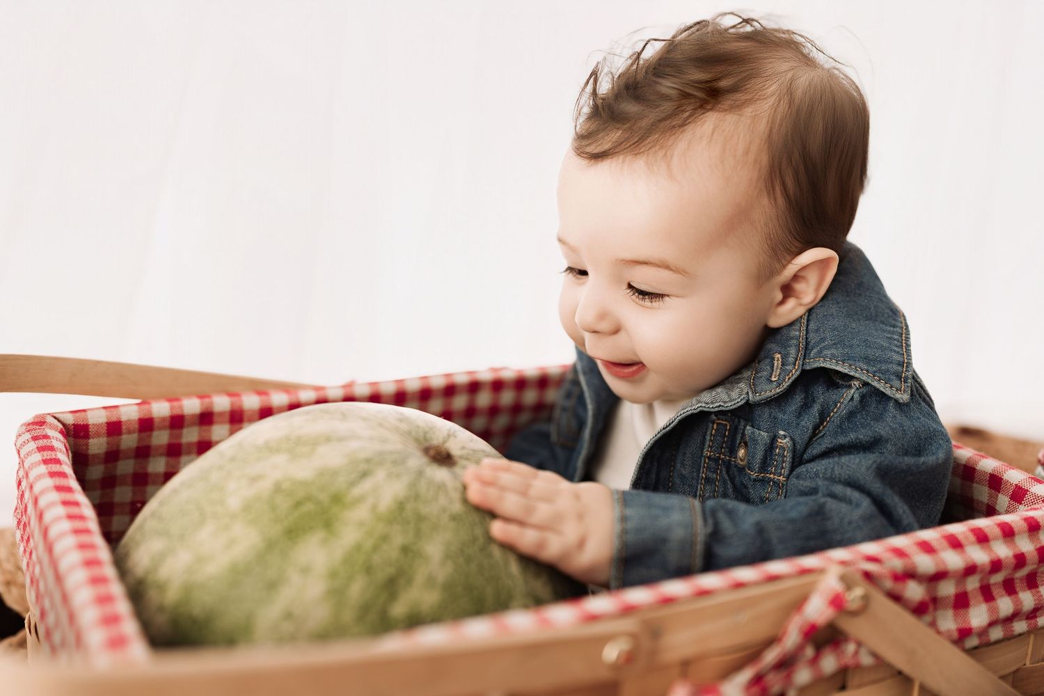 Adorable baby in denim jacket sitting in wooden crate exploring a green watermelon.
