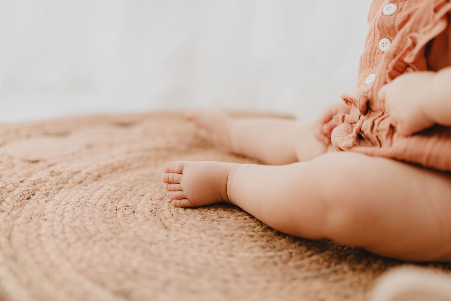 Close-up of baby feet and legs in rust romper sitting on woven jute rug.