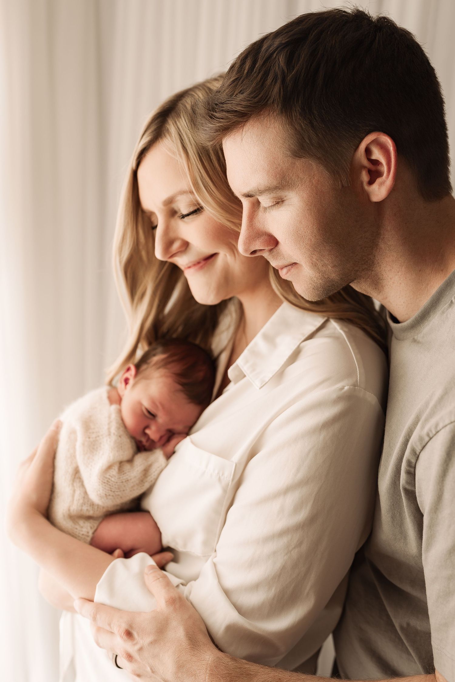 New parents smiling peacefully while holding their sleeping newborn baby in a bright studio.