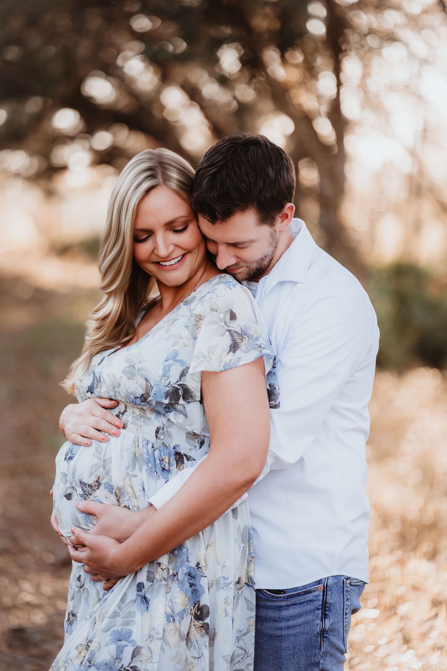 Man embracing pregnant woman in floral dress from behind in warm outdoor setting.