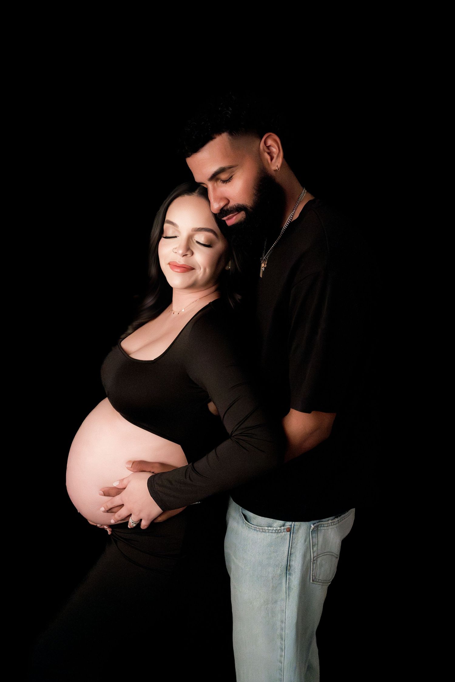 Couple embracing during maternity photo shoot on black background, man tenderly holding pregnant woman's belly.