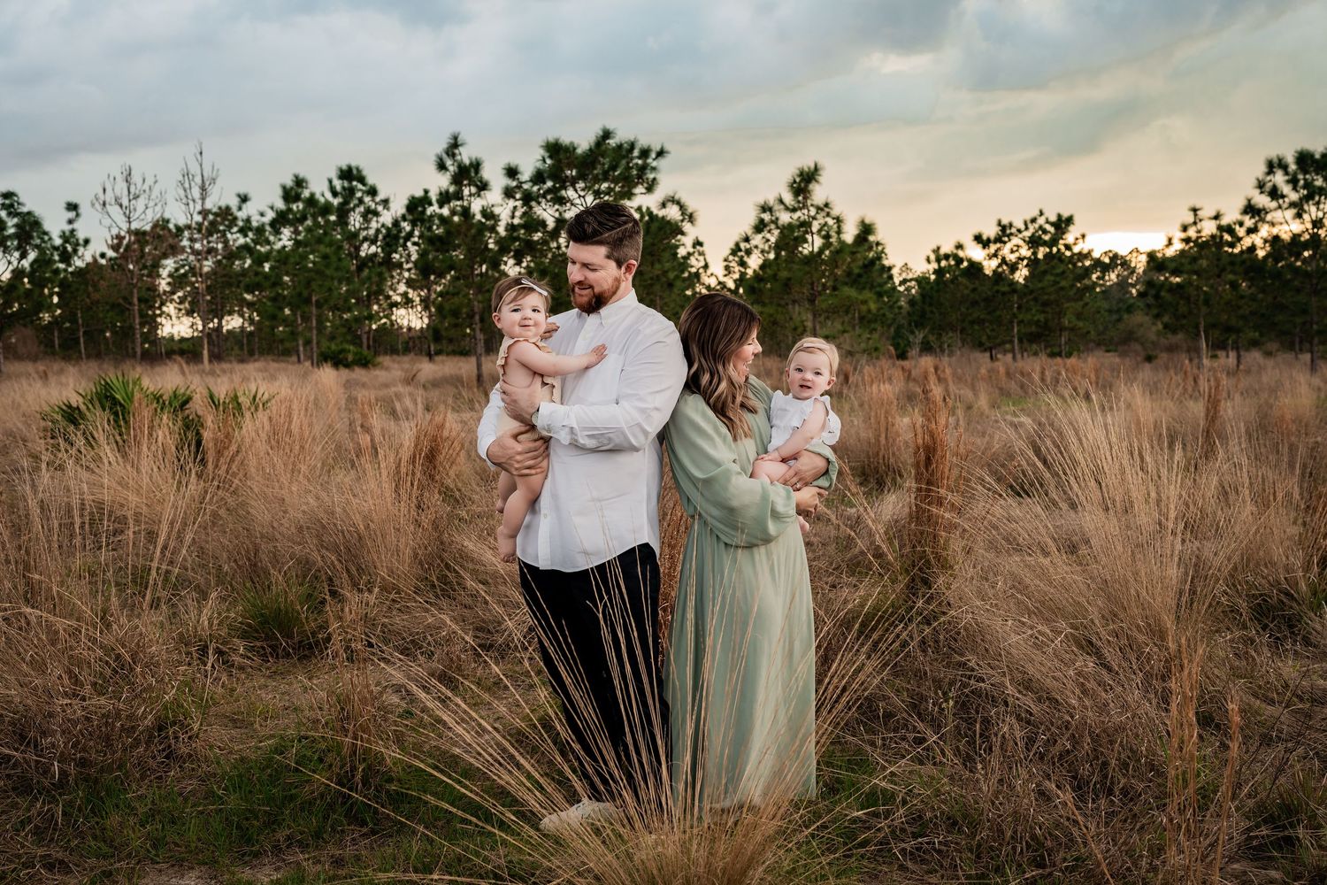 Family of four standing in tall golden grass field at dusk, parents holding two young babies.