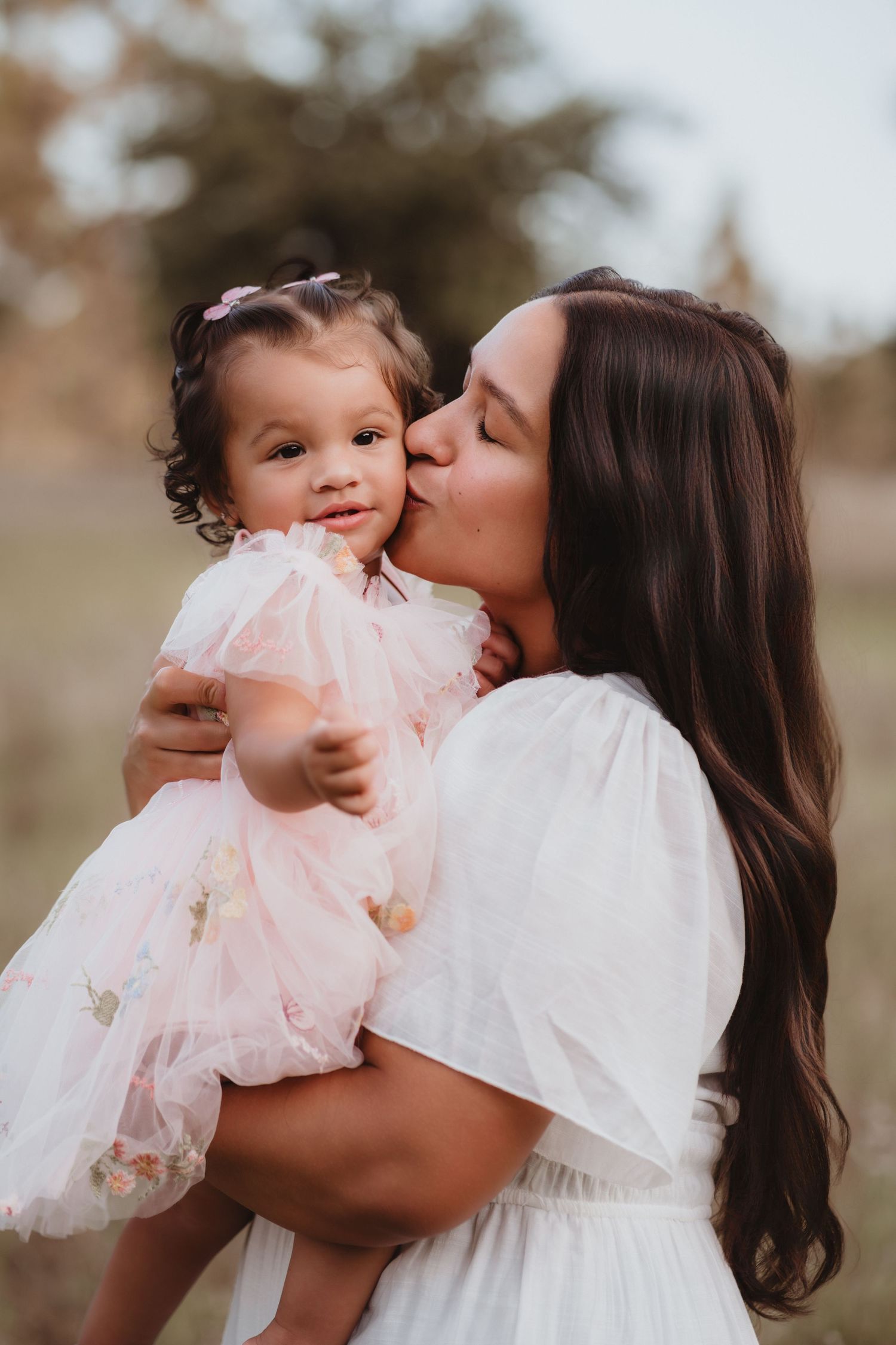 Loving mother in white dress kissing toddler daughter in pink tulle dress outside.