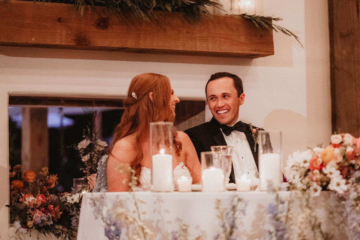 Bride with red hair and groom in black tuxedo laugh together at floral-decorated sweetheart table.