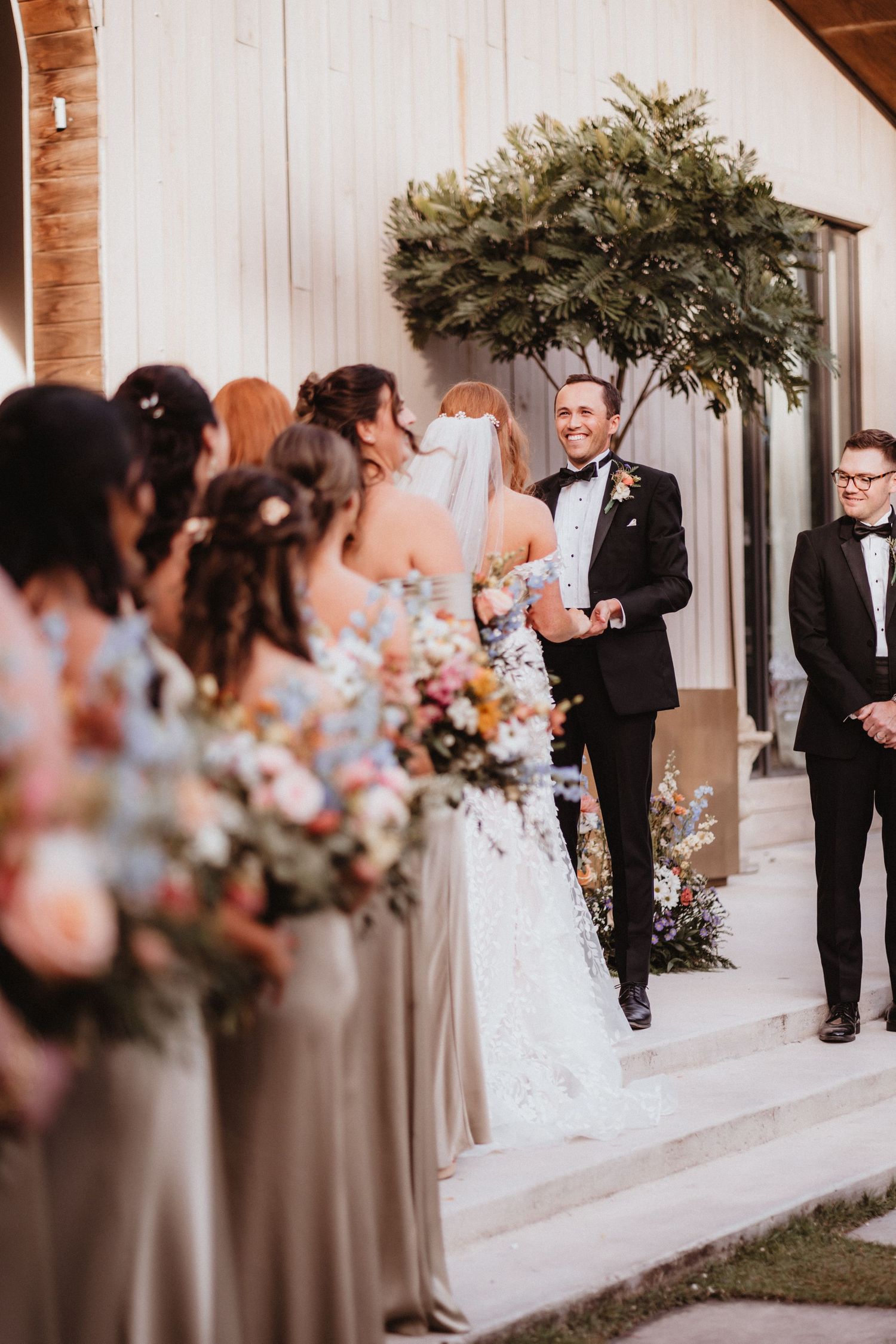 Groom in black tuxedo smiles at bride arriving at altar with bridesmaids holding bouquets nearby.