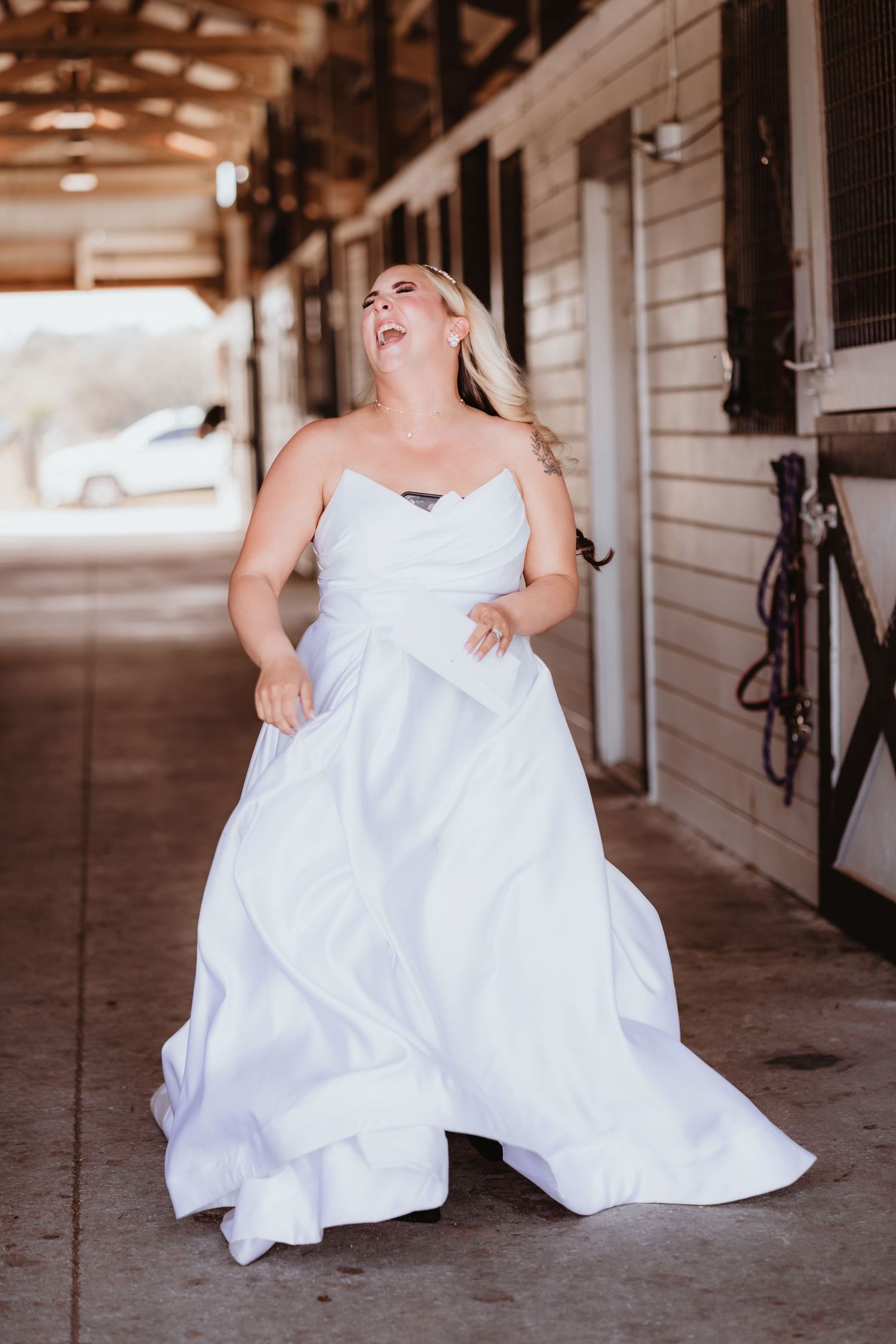 Radiant bride in strapless white wedding gown laughs joyfully while walking through a sunlit rustic barn.