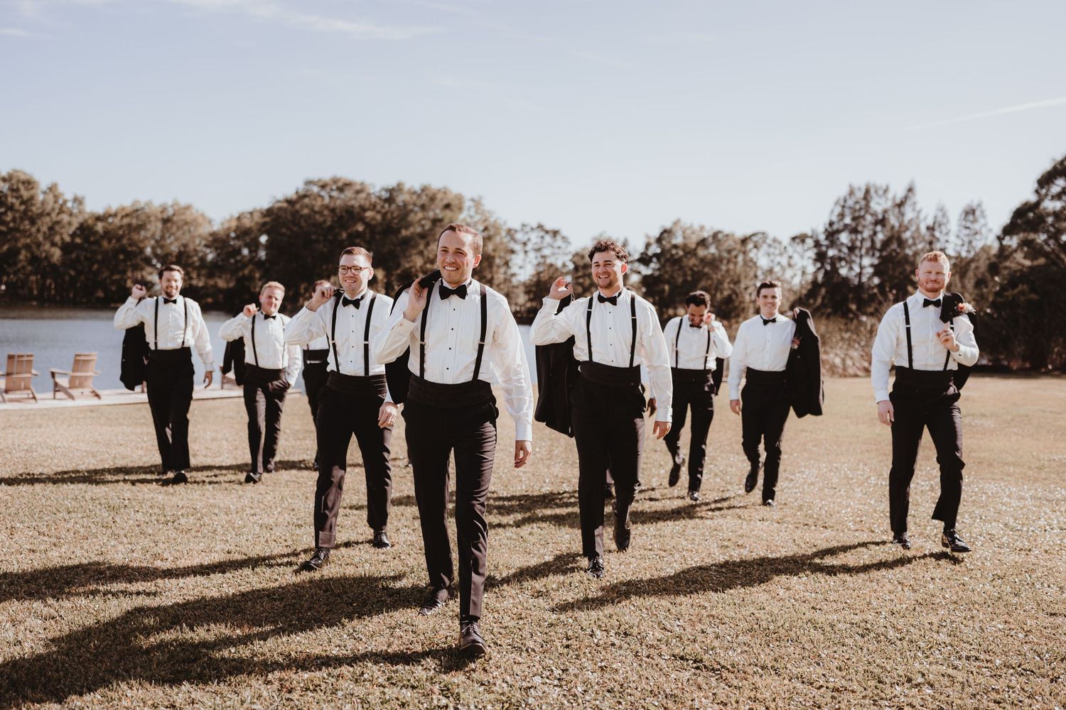 Groomsmen in black tuxedos with suspenders walking confidently across a green lawn near a lake.