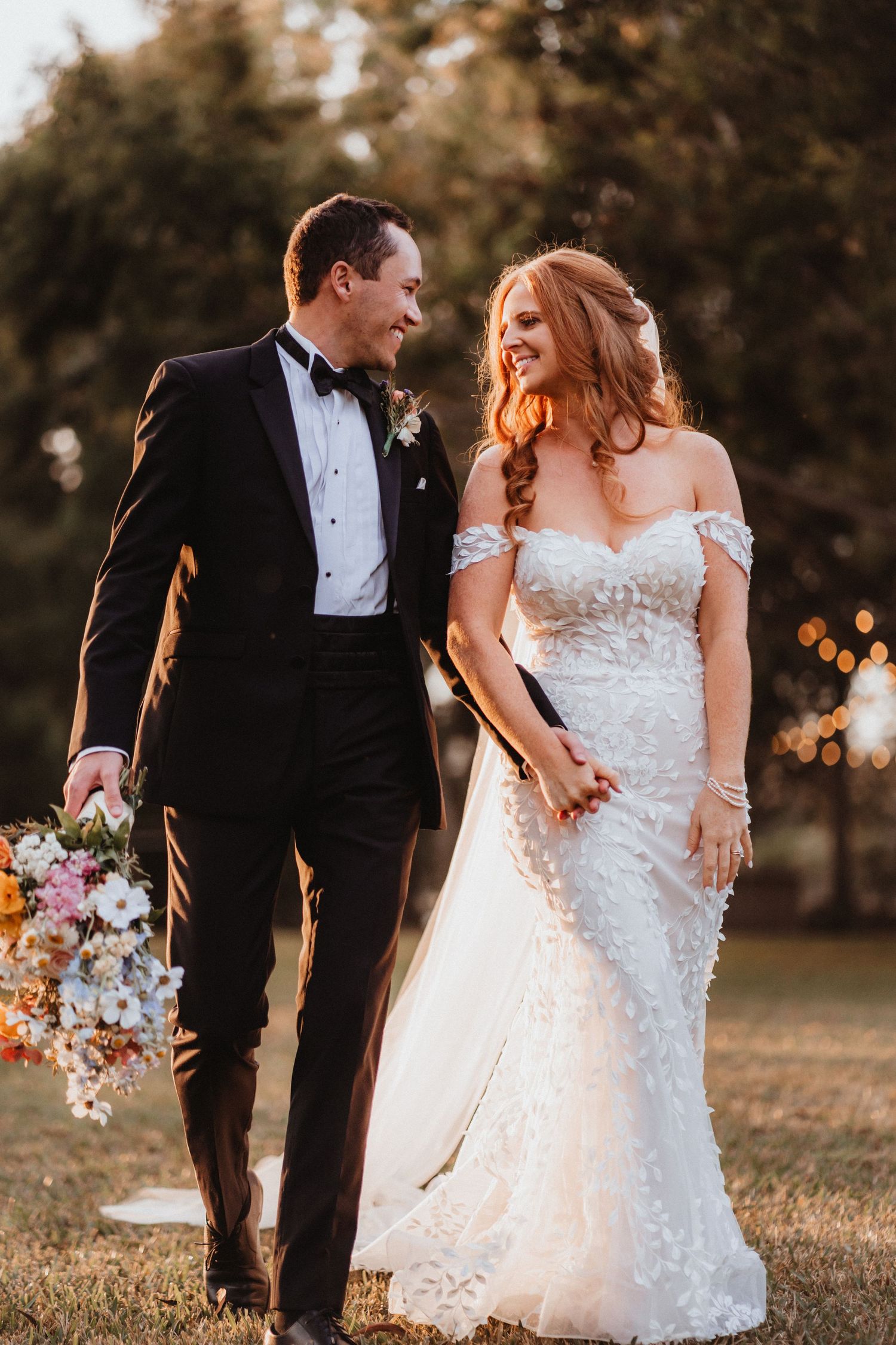 Elegant bride and groom stroll hand in hand at sunset, she in lace gown with veil, he in black bow tie tuxedo.