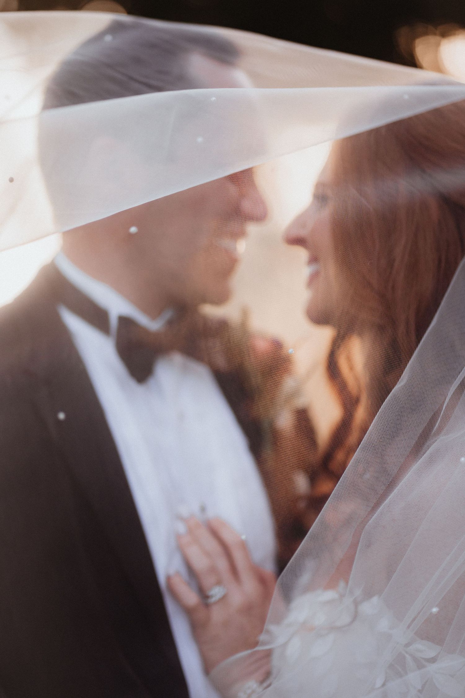 Romantic soft-focus portrait of bride and groom smiling under a sheer veil in golden backlit light.