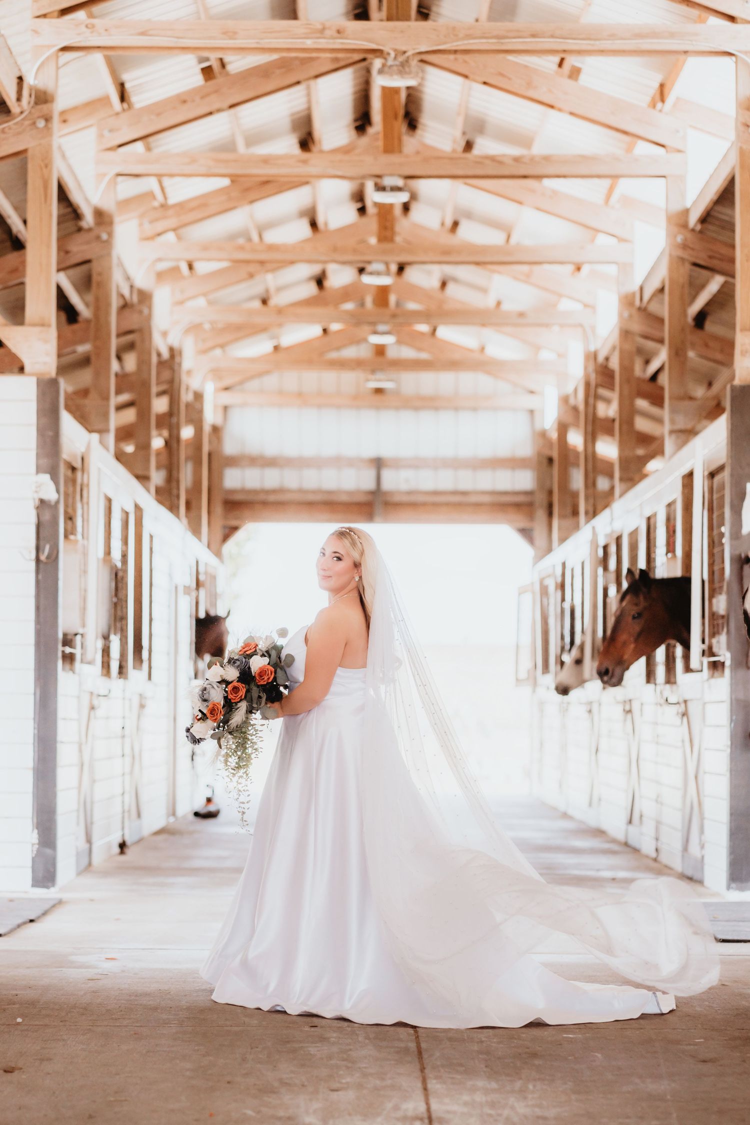 Bride in a white gown and veil stands in a sunlit horse stable holding a bouquet of orange and white roses.