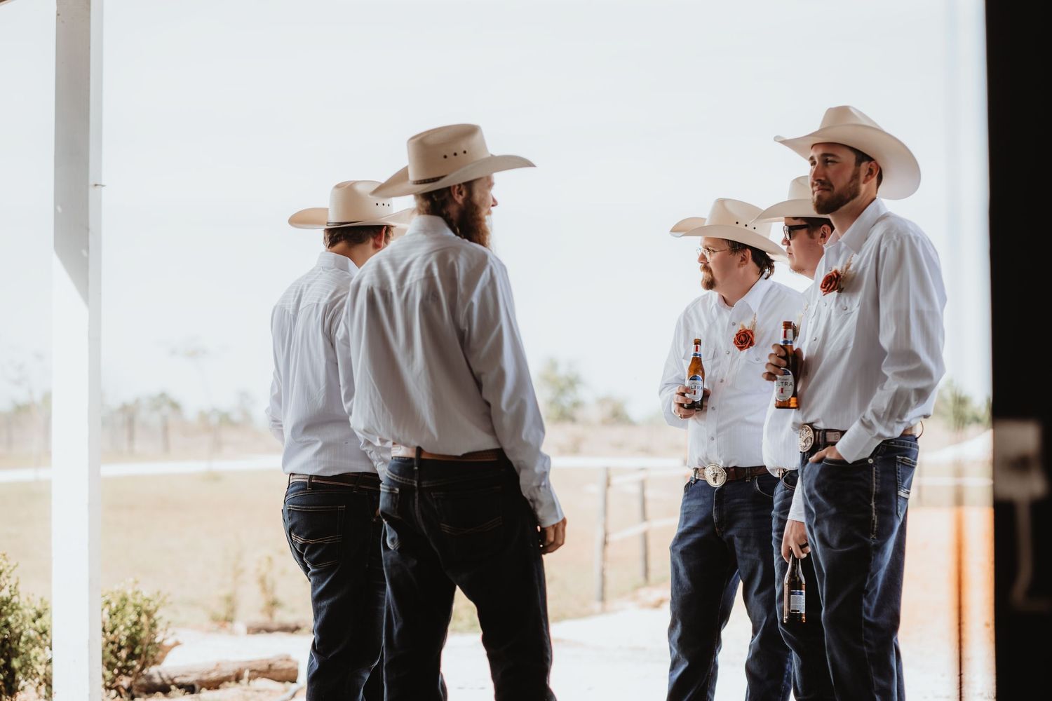 Groomsmen in white shirts, jeans, and cowboy hats gather under a barn porch at a rustic wedding.