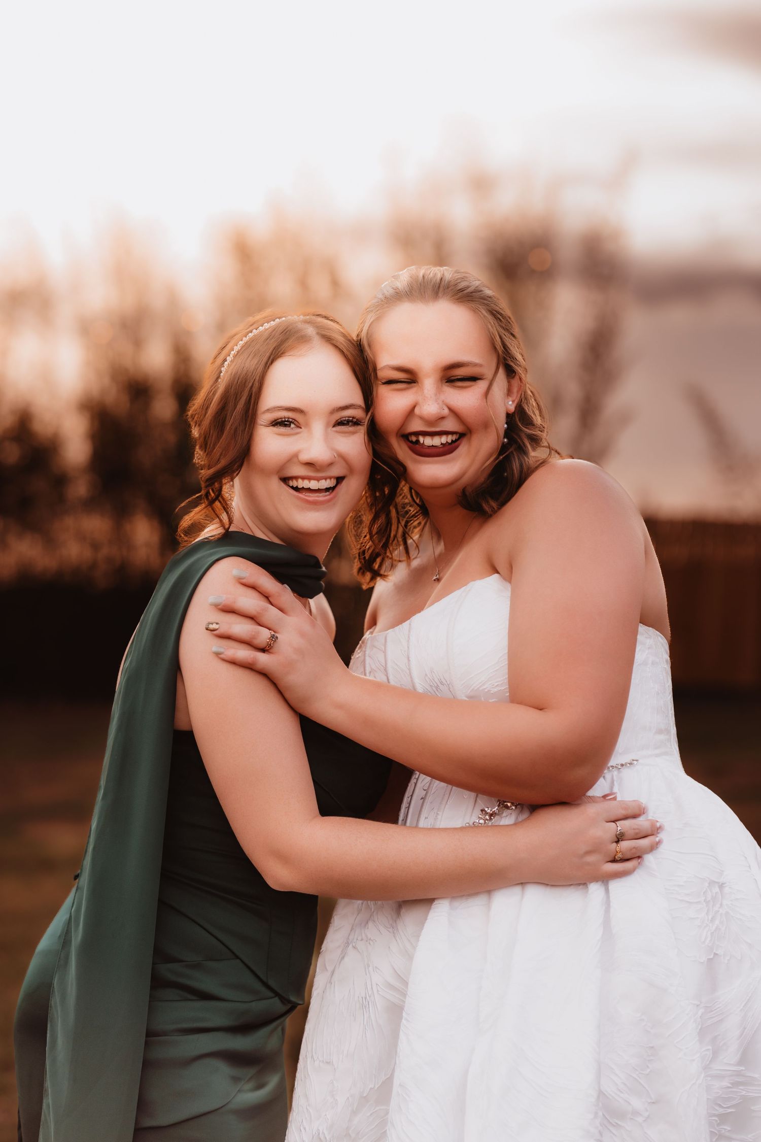 Smiling bride in white lace gown hugs bridesmaid in green dress at golden hour.