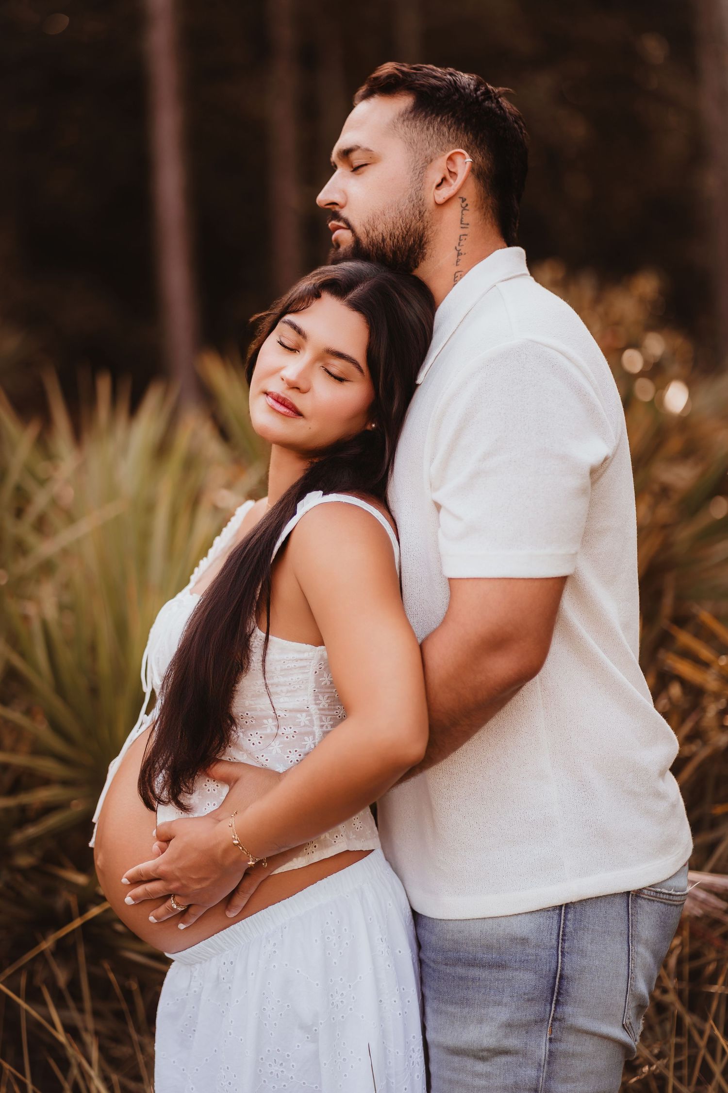Expecting couple in white outfits embracing tenderly outdoors, man holding pregnant womans belly from behind.
