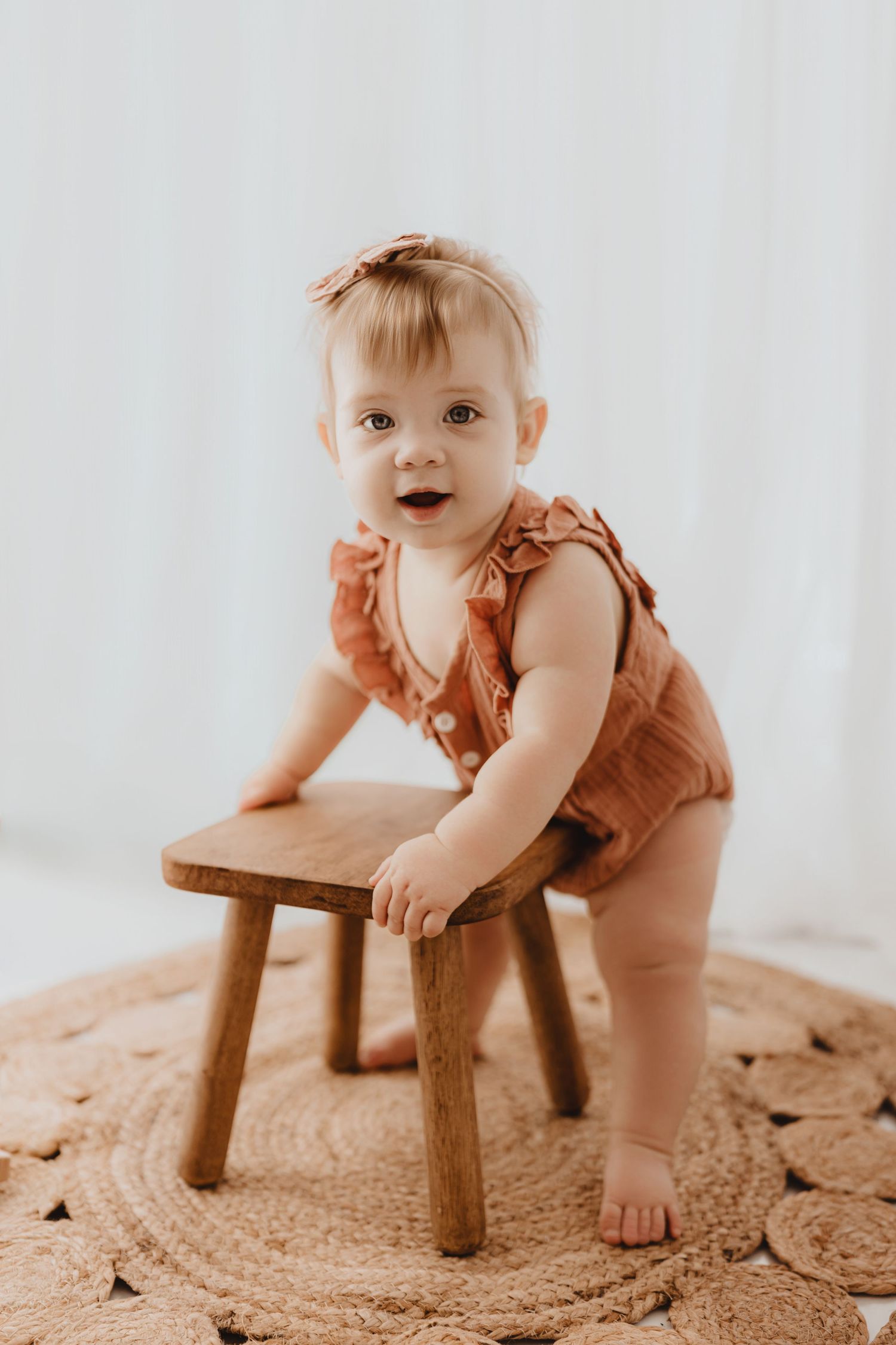 Baby girl in terracotta ruffle romper standing at a small wooden stool on a woven rug.