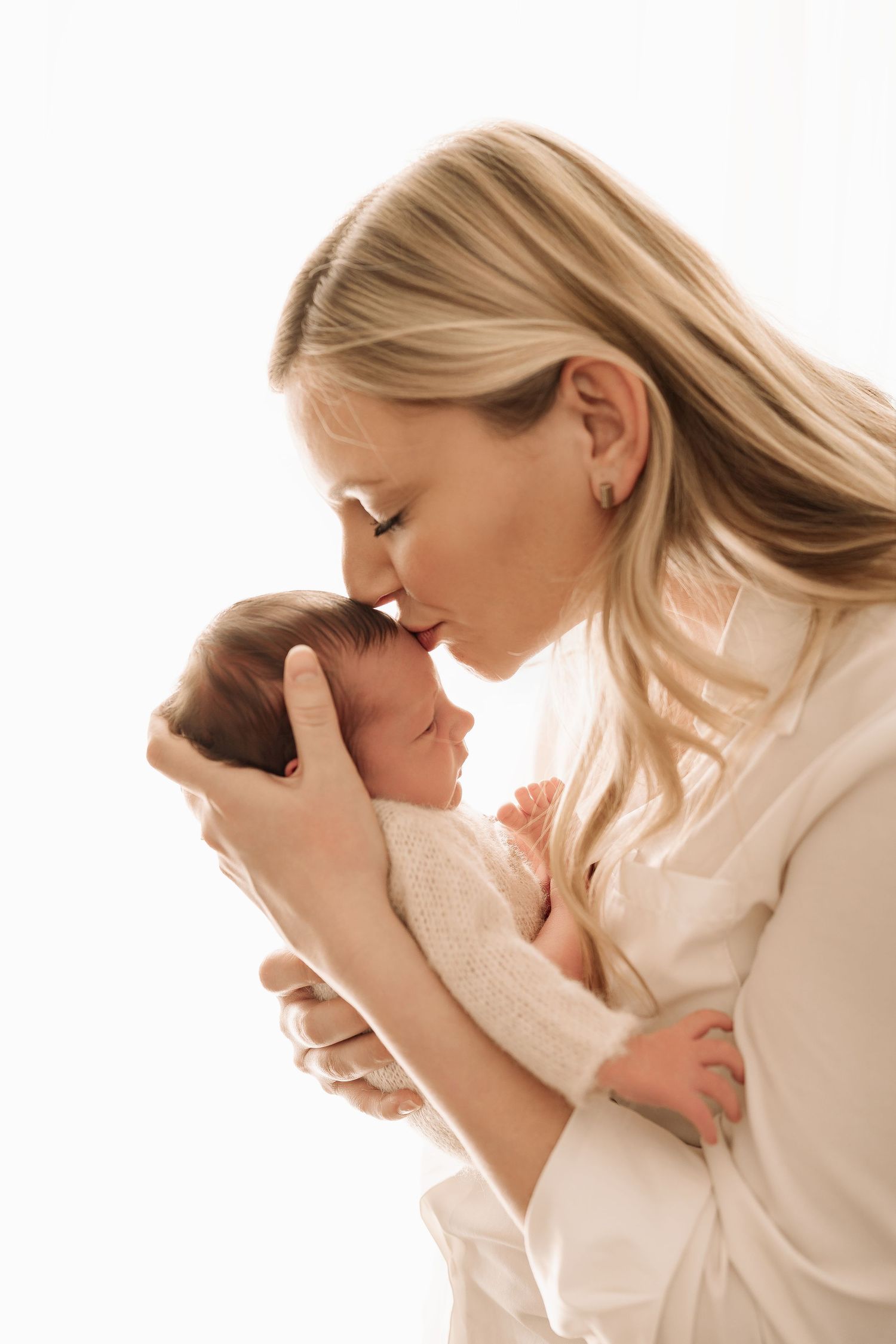 Blonde mother kissing forehead of sleeping newborn baby wrapped in cream knit swaddle.