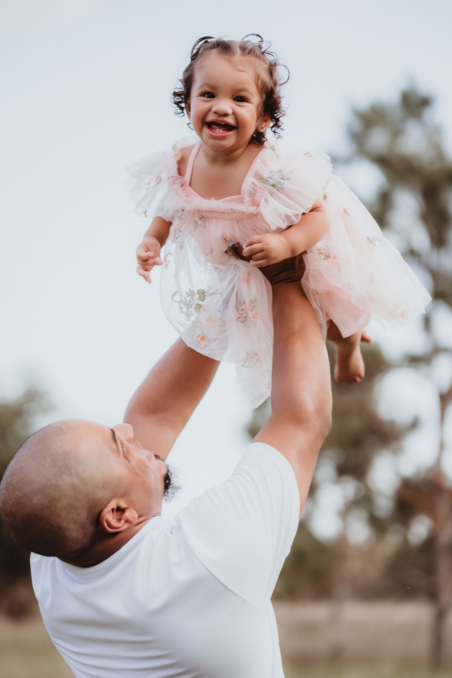Playful father lifting his giggling toddler daughter in a pink tulle dress high in the air.