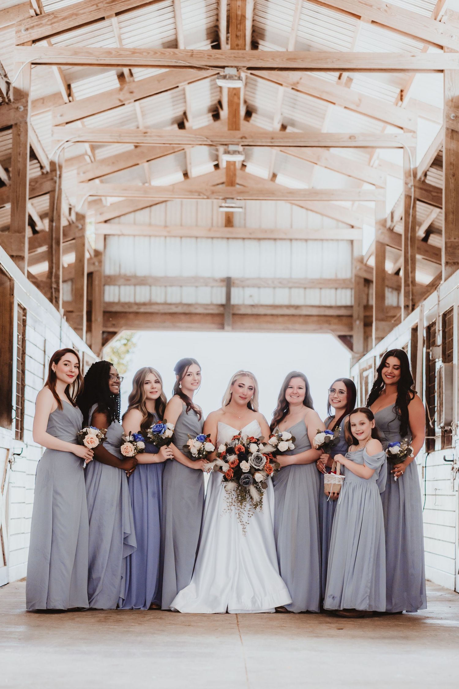 Bride and bridesmaids in dusty blue gowns pose with bouquets inside a wooden barn venue.