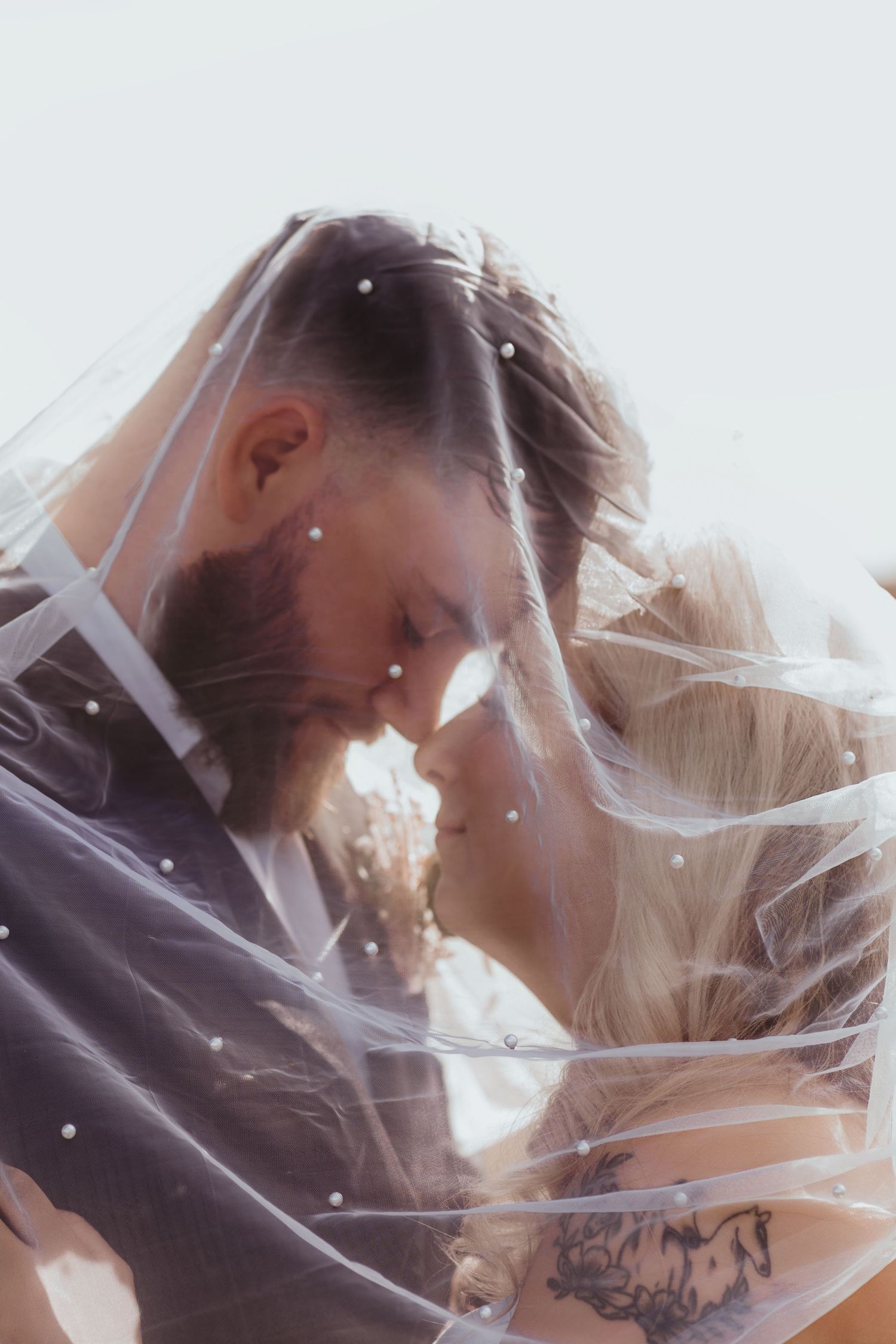 Bride and groom share an intimate forehead touch under a pearl-adorned wedding veil in a dreamy portrait.