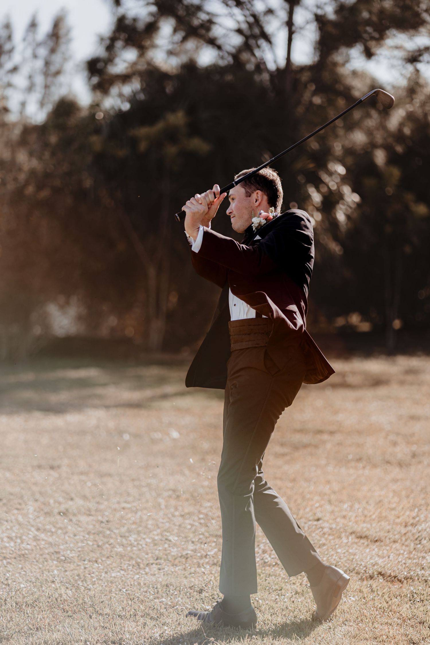 Groom in burgundy tuxedo jacket swings a golf club on a sunny field before his wedding.