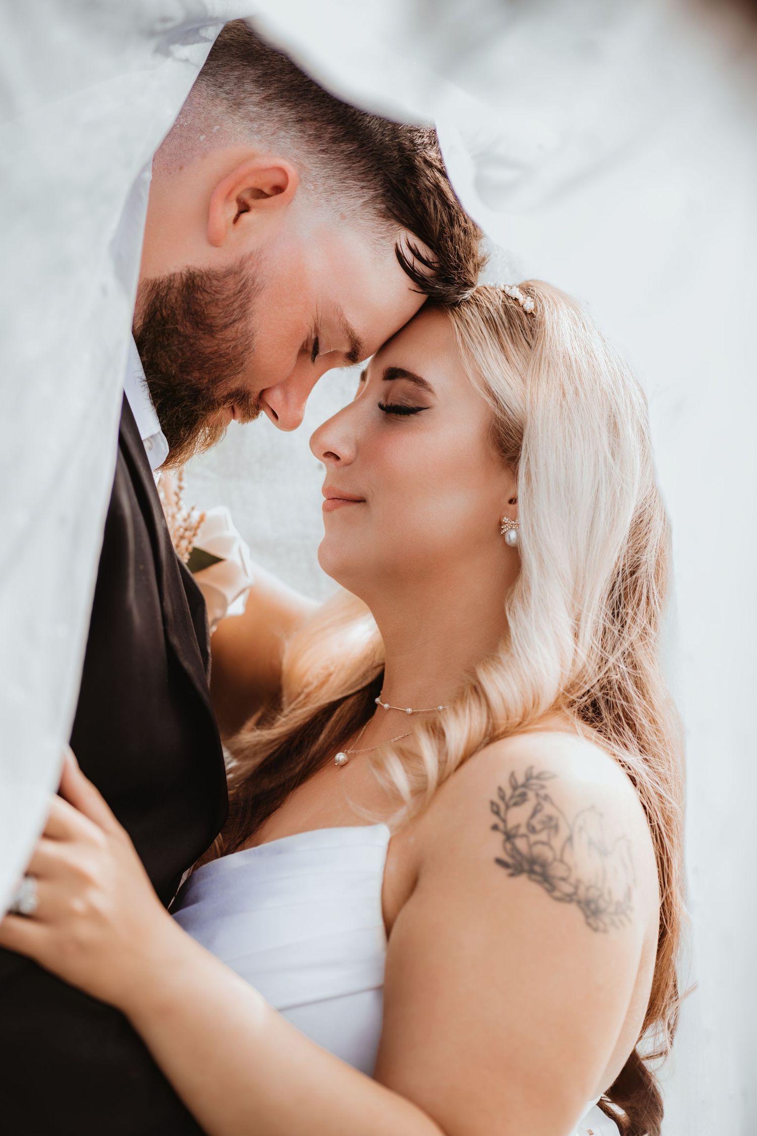 Bride and groom touch foreheads tenderly under a veil, bride wearing pearl earrings and a floral shoulder tattoo.