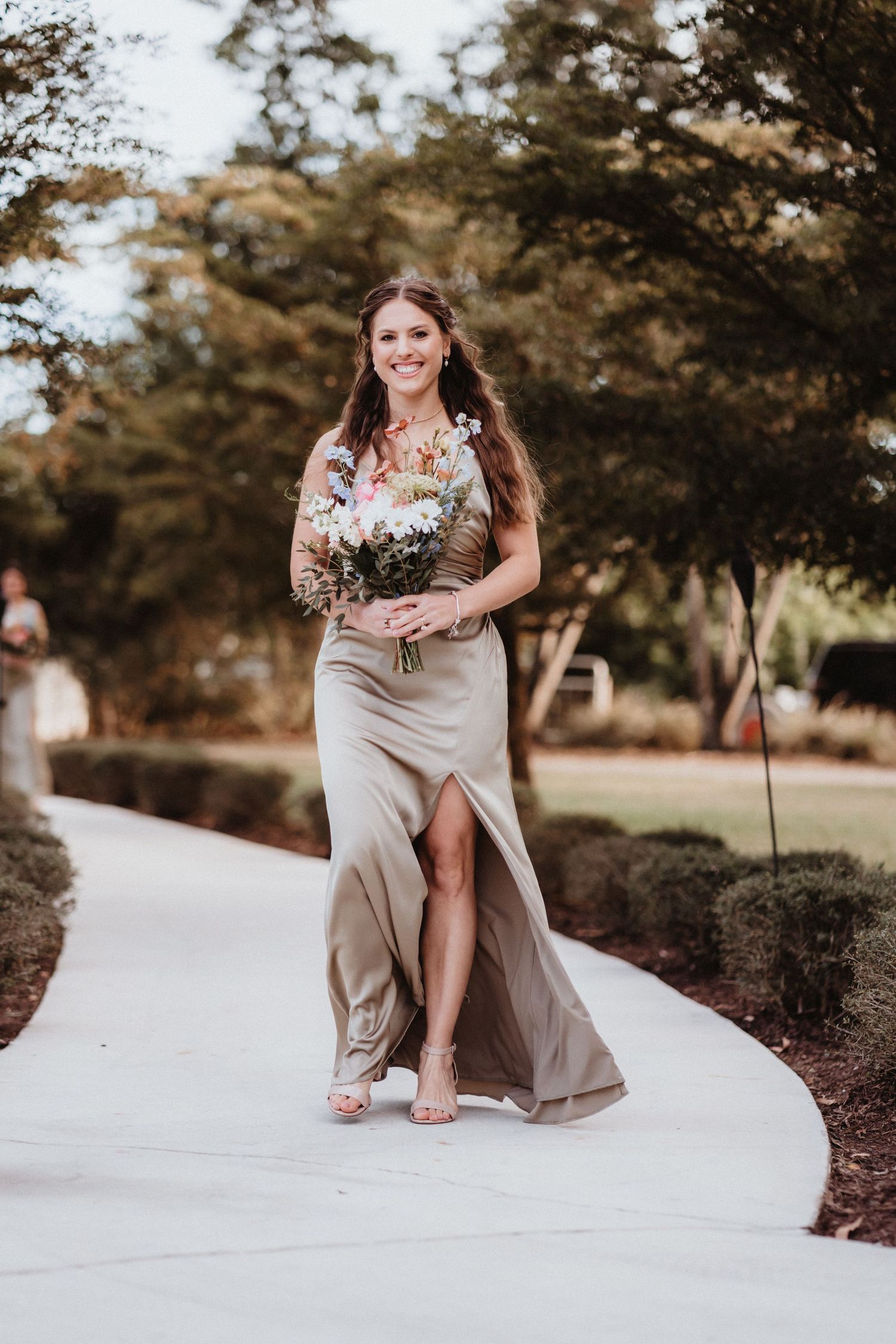 Smiling bridesmaid in sage green satin gown carrying a wildflower bouquet down the aisle.