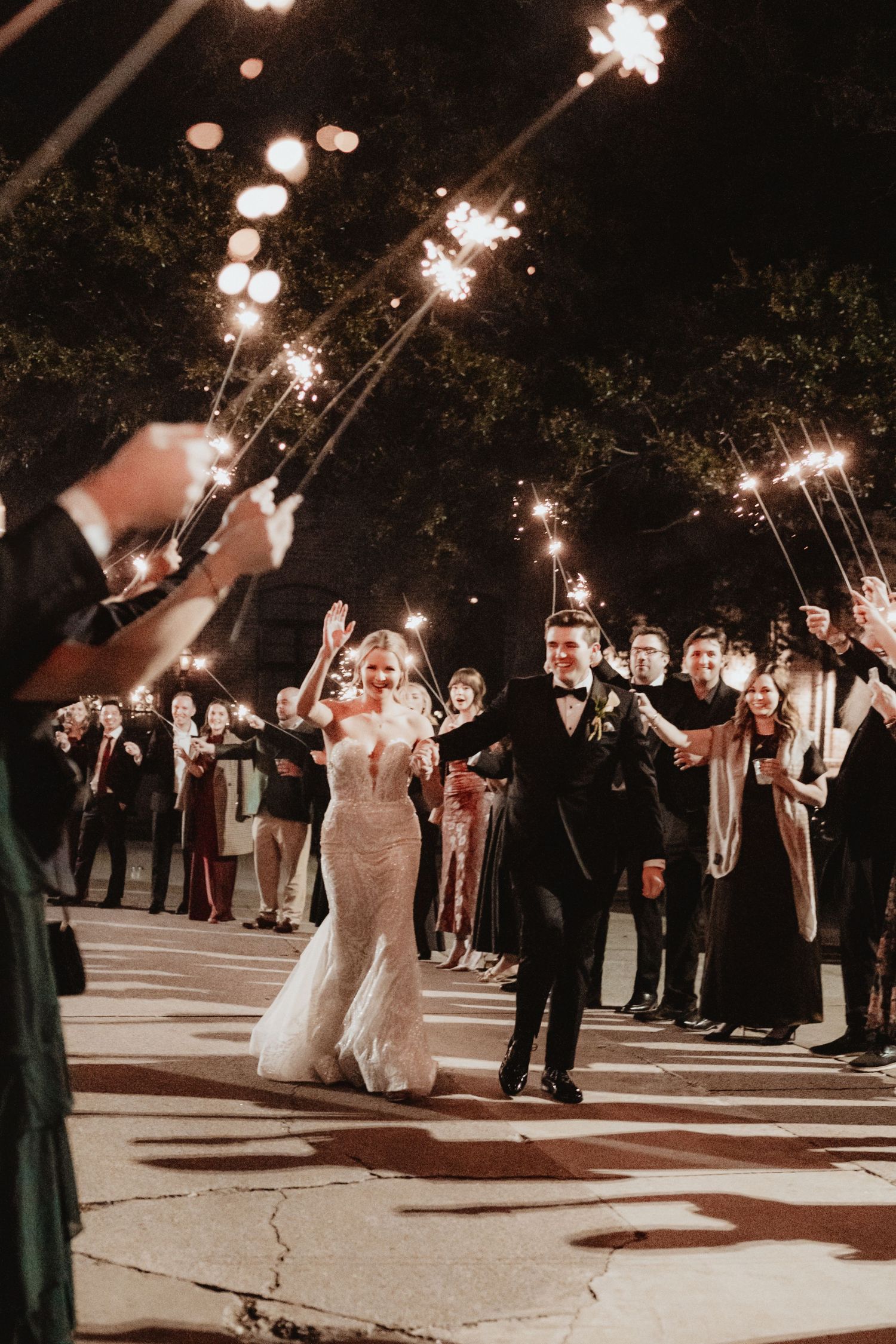 Bride and groom exit through a glittering sparkler tunnel surrounded by cheering wedding guests at night.