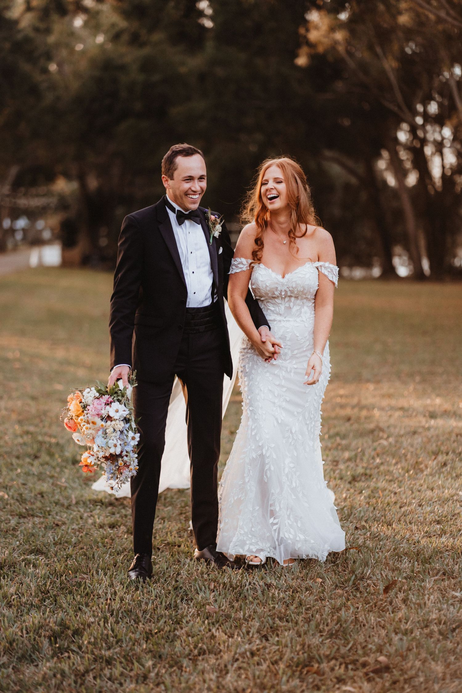 Laughing newlyweds walk hand in hand across green lawn, groom in black tux, bride in off-shoulder lace wedding gown.