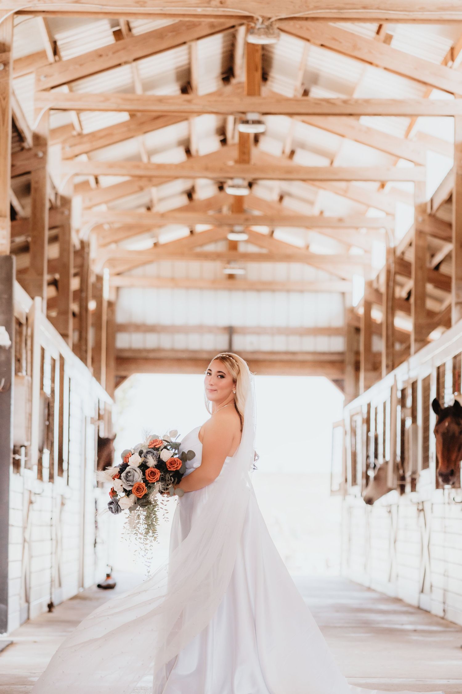Bride in a white gown and veil glances back while holding a bouquet in a warmly lit horse stable.