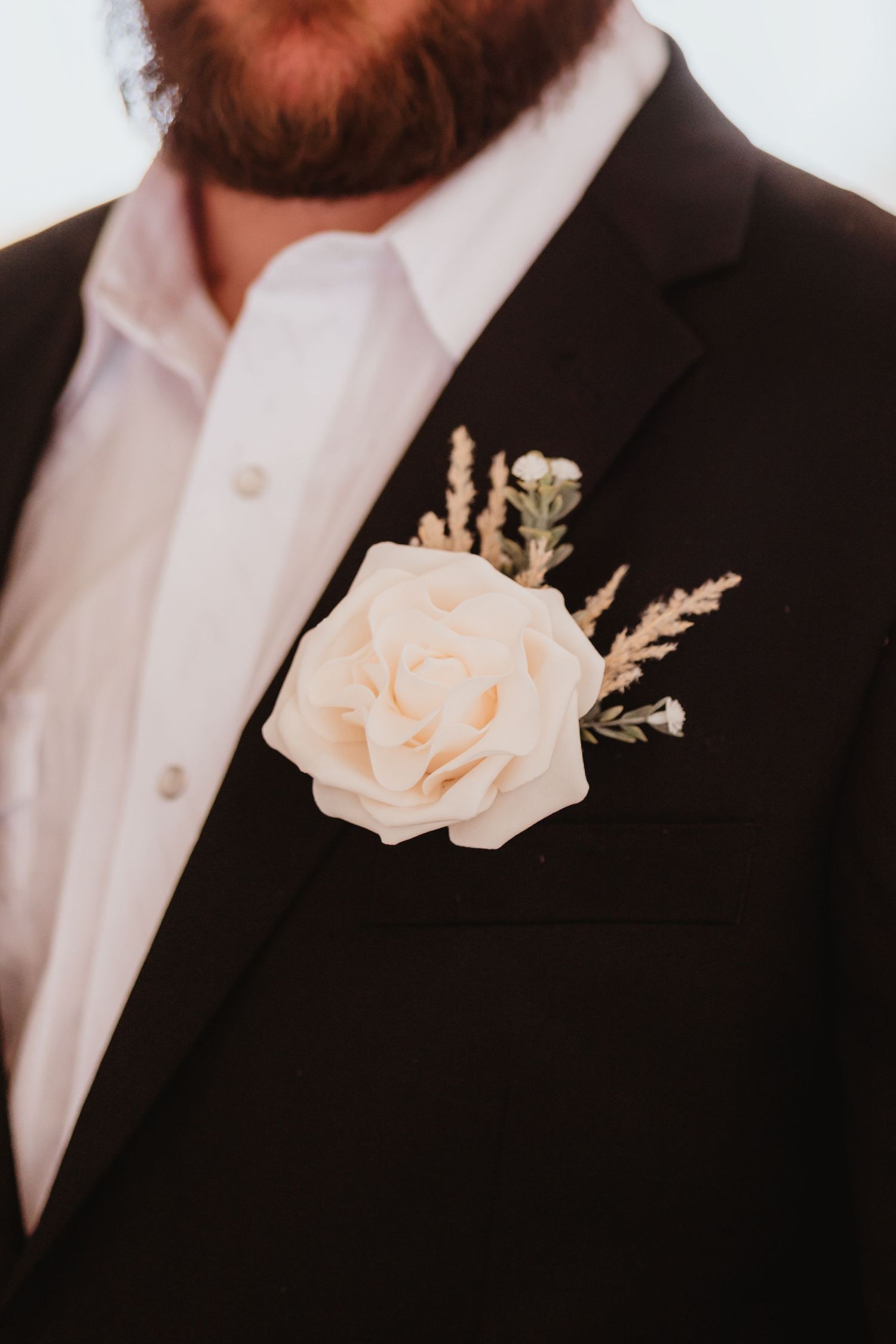 Close-up of white rose boutonniere with greenery on groom's black wedding suit lapel.