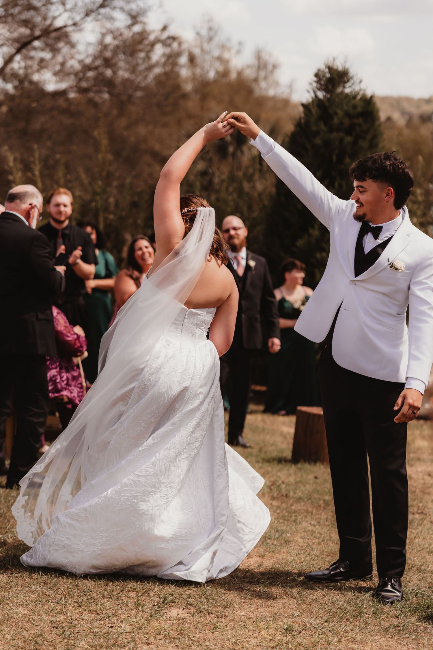 Groom in white tuxedo twirling bride in flowing white wedding gown during outdoor ceremony recessional.