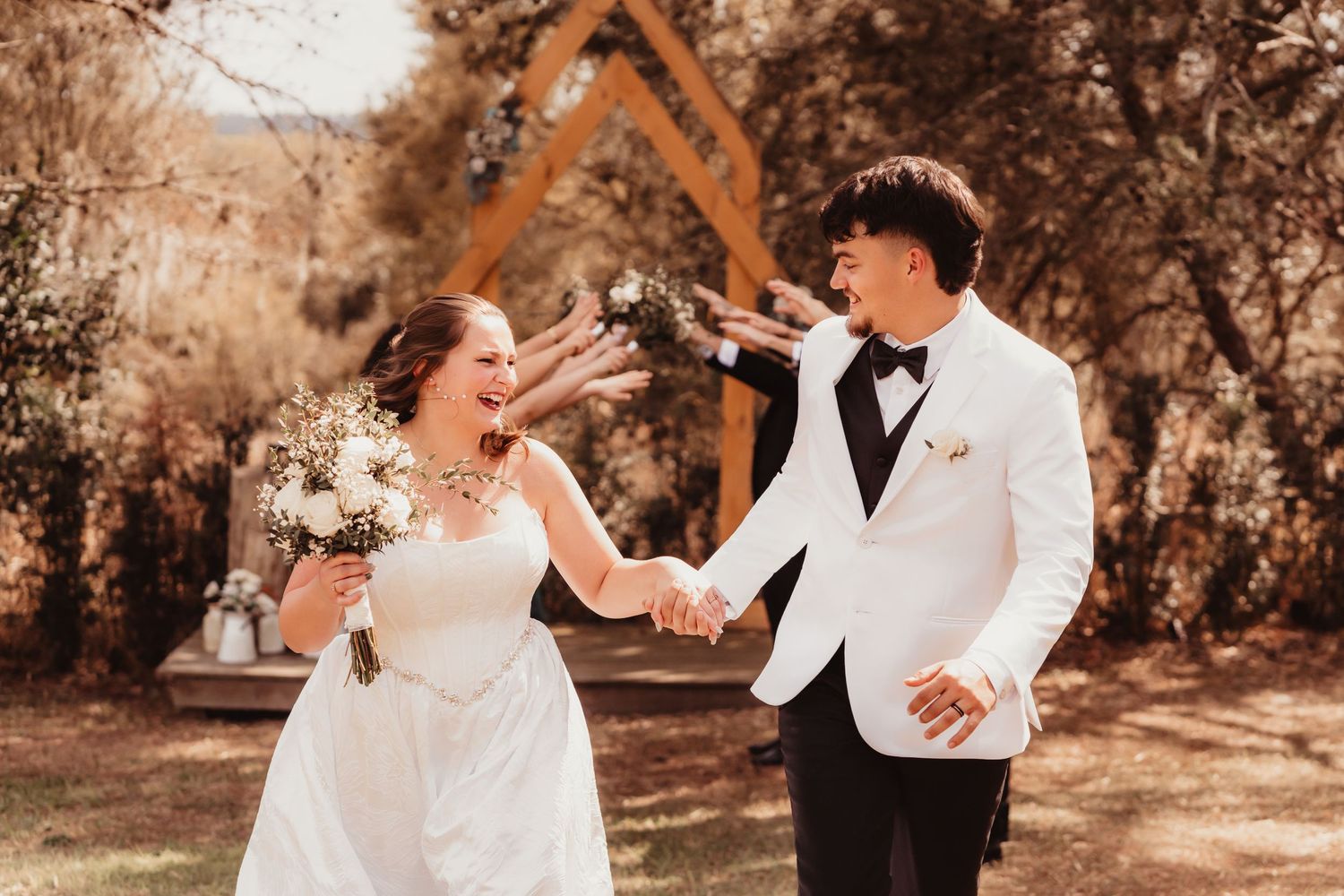 Happy bride and groom holding hands and laughing together at outdoor rustic wedding venue.