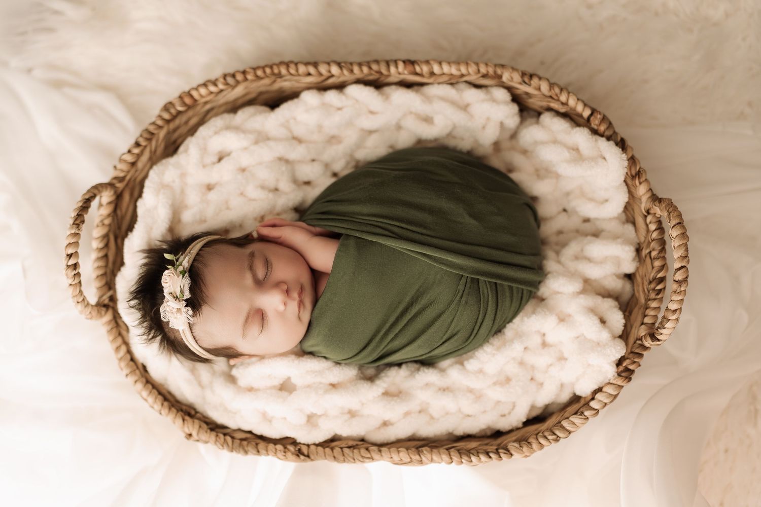Newborn baby swaddled in olive green wrap sleeping in wicker basket with chunky knit blanket.