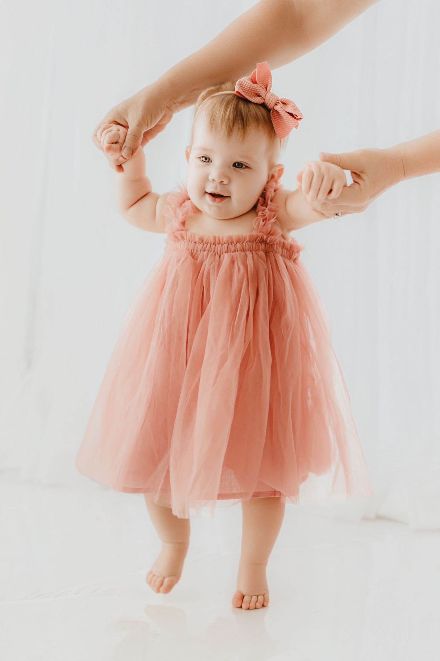 Baby girl in coral tulle dress and pink bow headband learning to walk with adult assistance in studio.