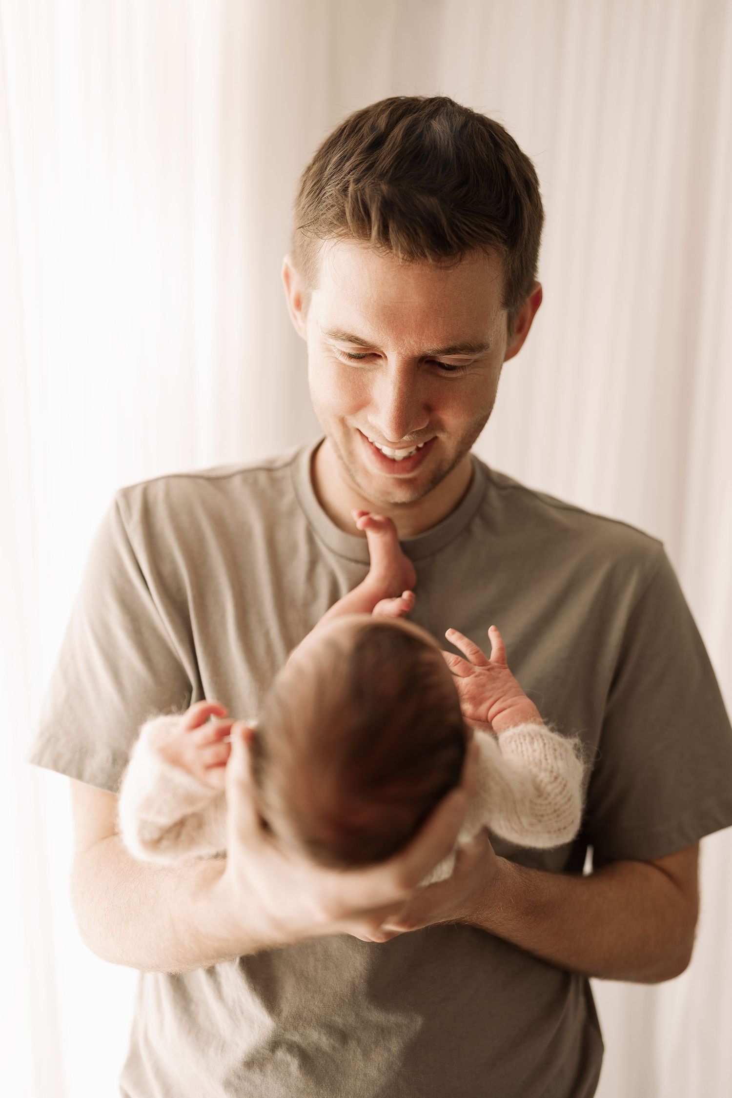 Smiling father holds newborn baby up while baby grabs his finger.