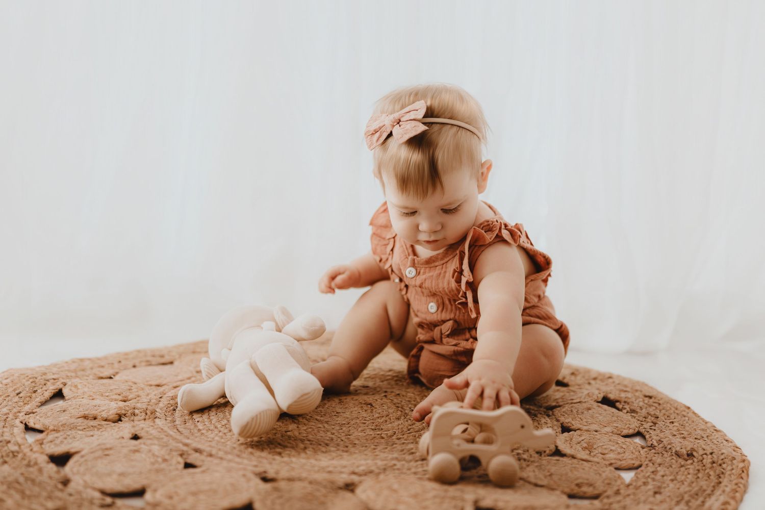 Baby girl in rust romper playing with wooden toys on round woven rug.