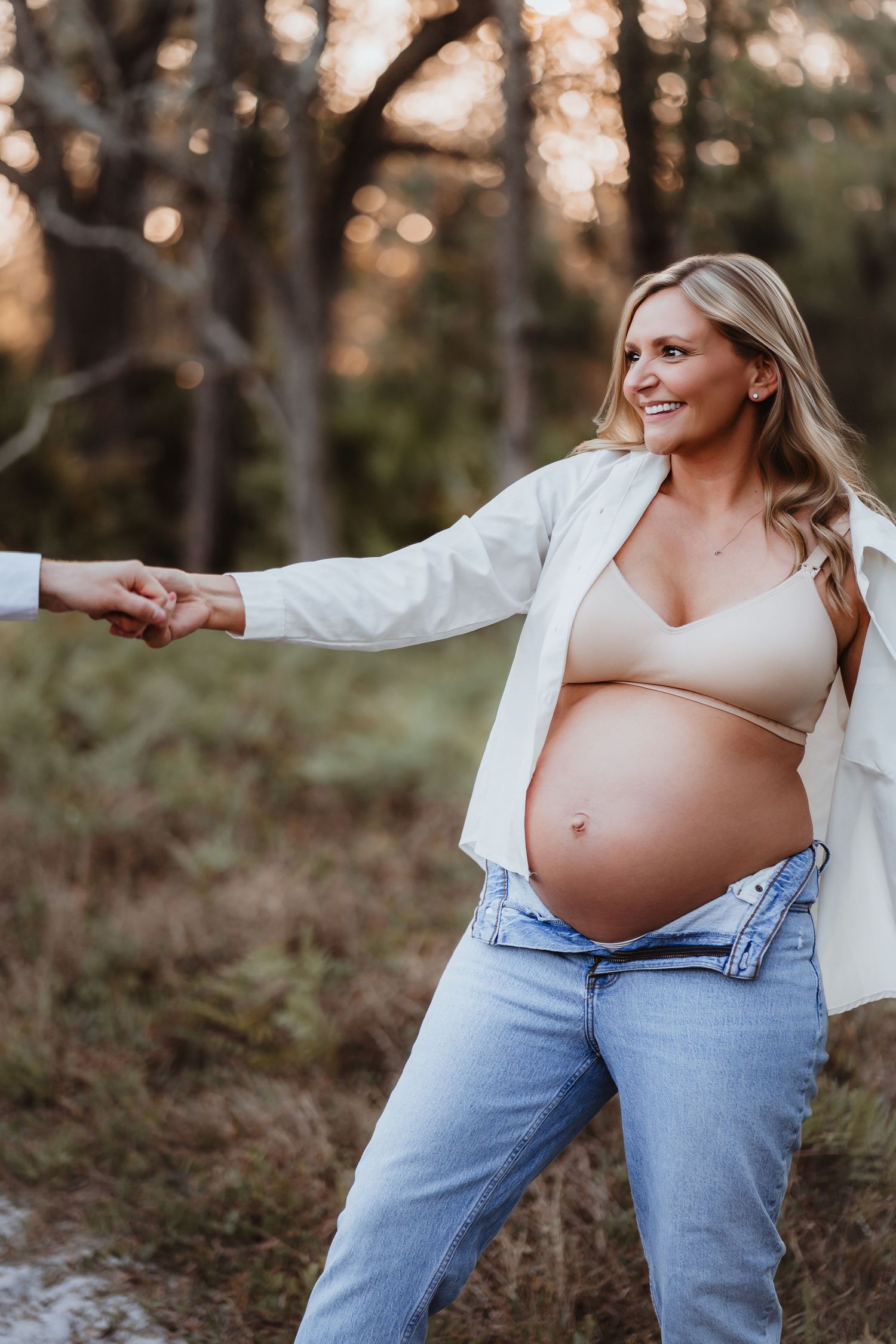 Smiling pregnant blonde woman holding partner's hand in a beautiful forest setting.