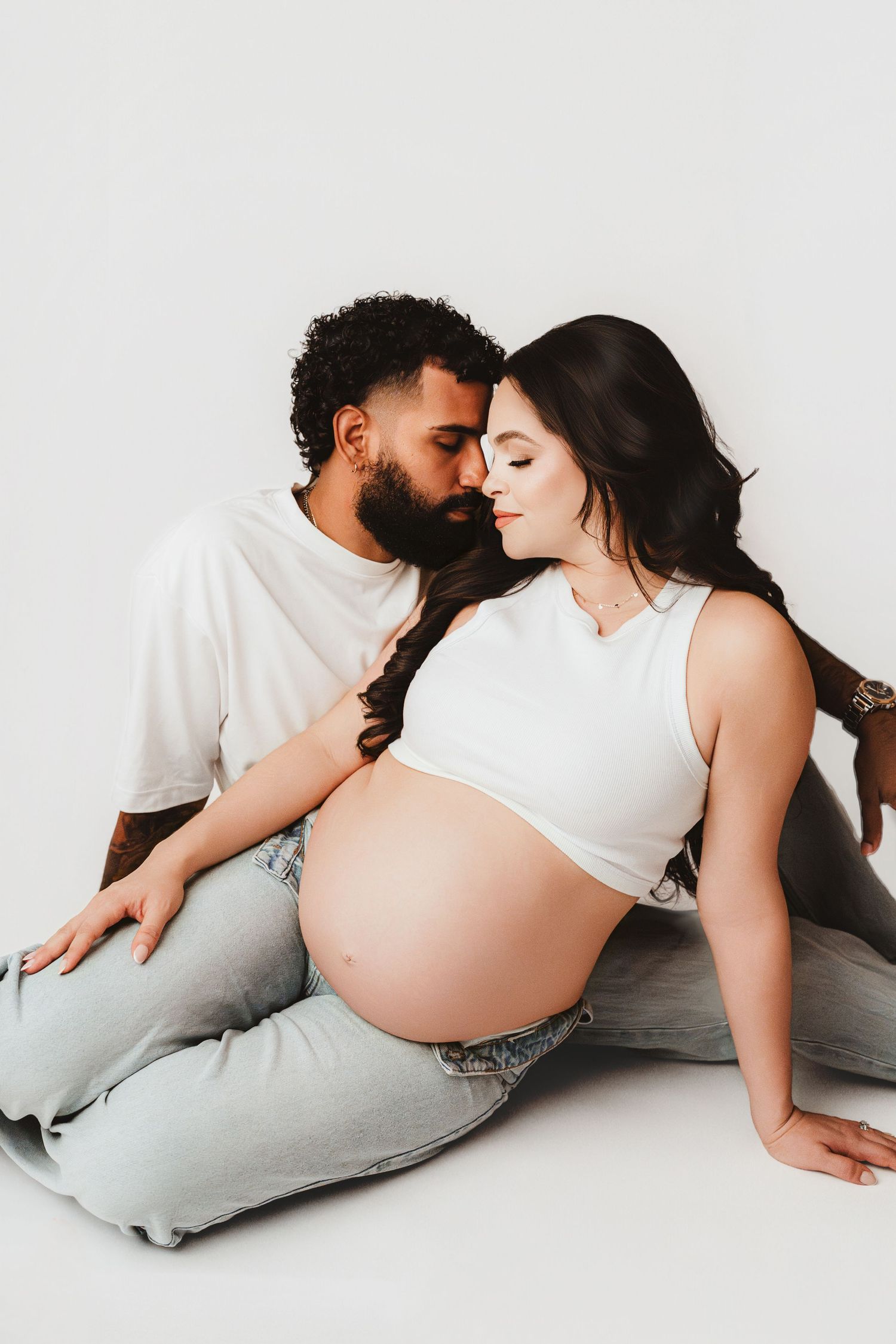 Couple sharing tender nose-to-nose moment, woman in white crop top showing pregnant belly.