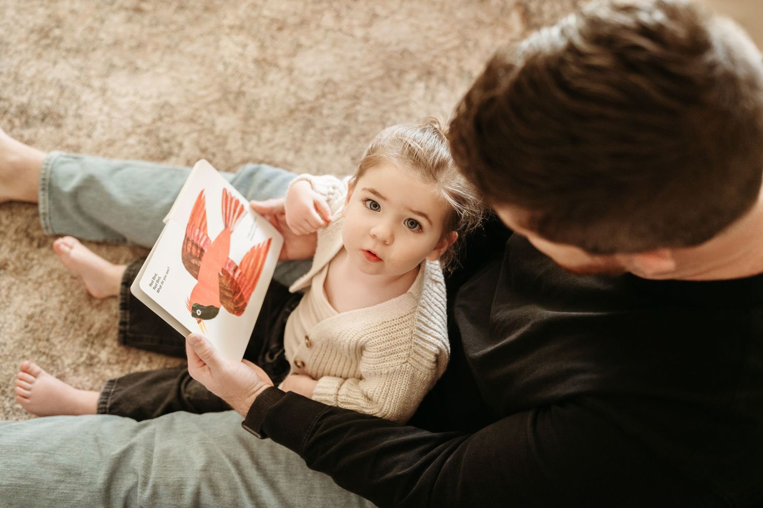 Father and toddler daughter reading a colorful picture book together while sitting on a cozy carpet floor.