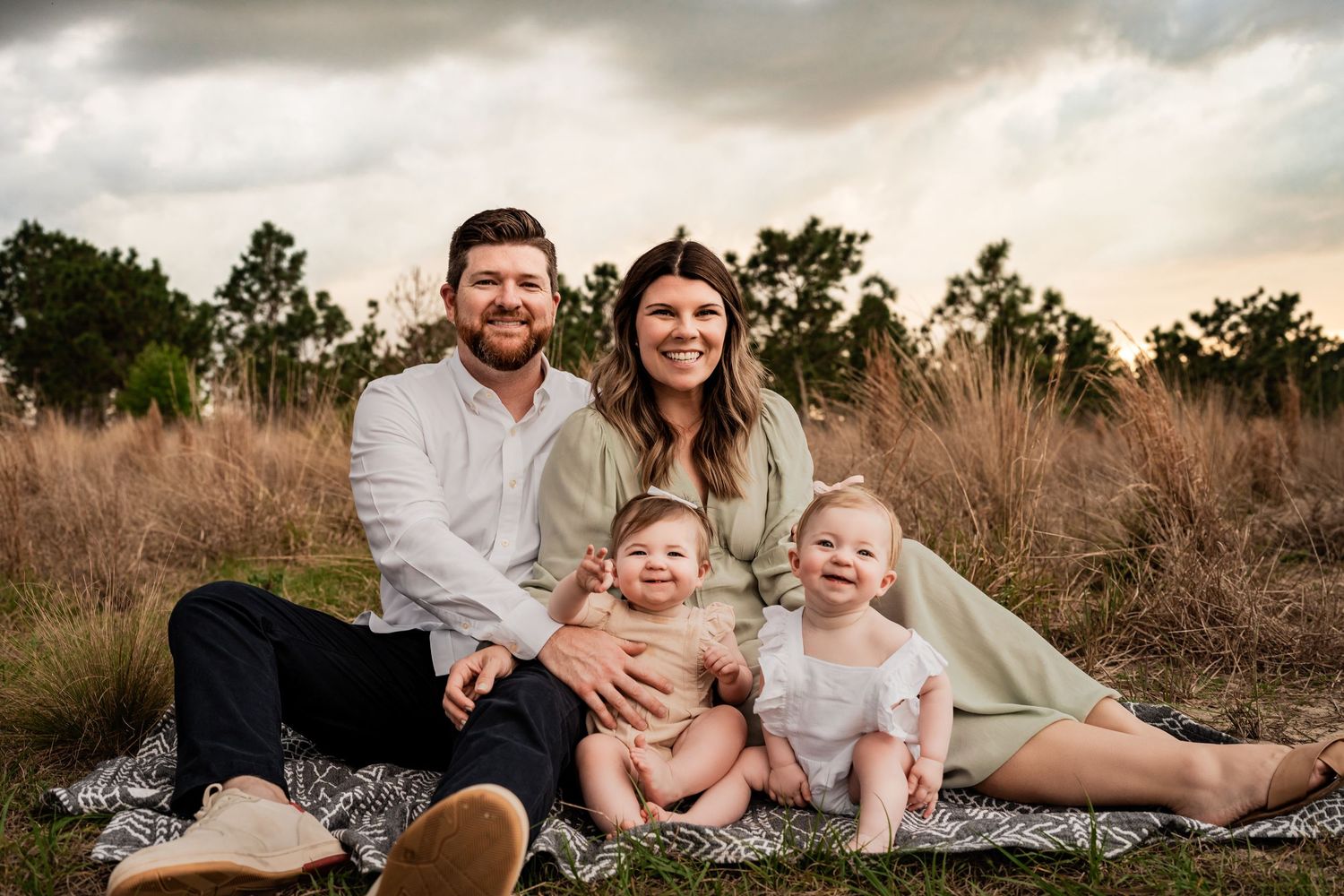 Happy family of four with two smiling baby girls sitting on a blanket in a golden sunset field.