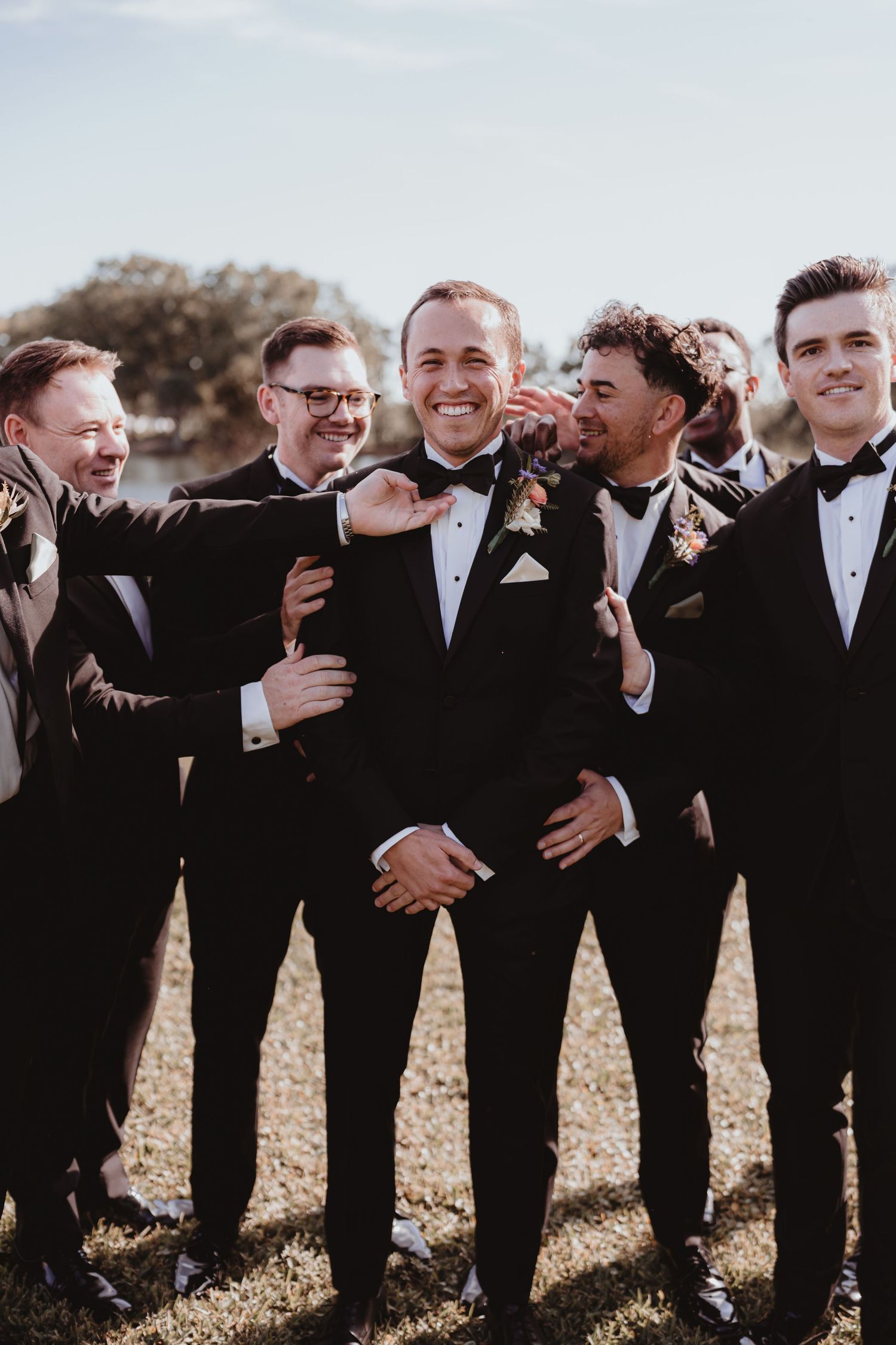 Groom laughing with groomsmen in matching black tuxedos outdoors at wedding.