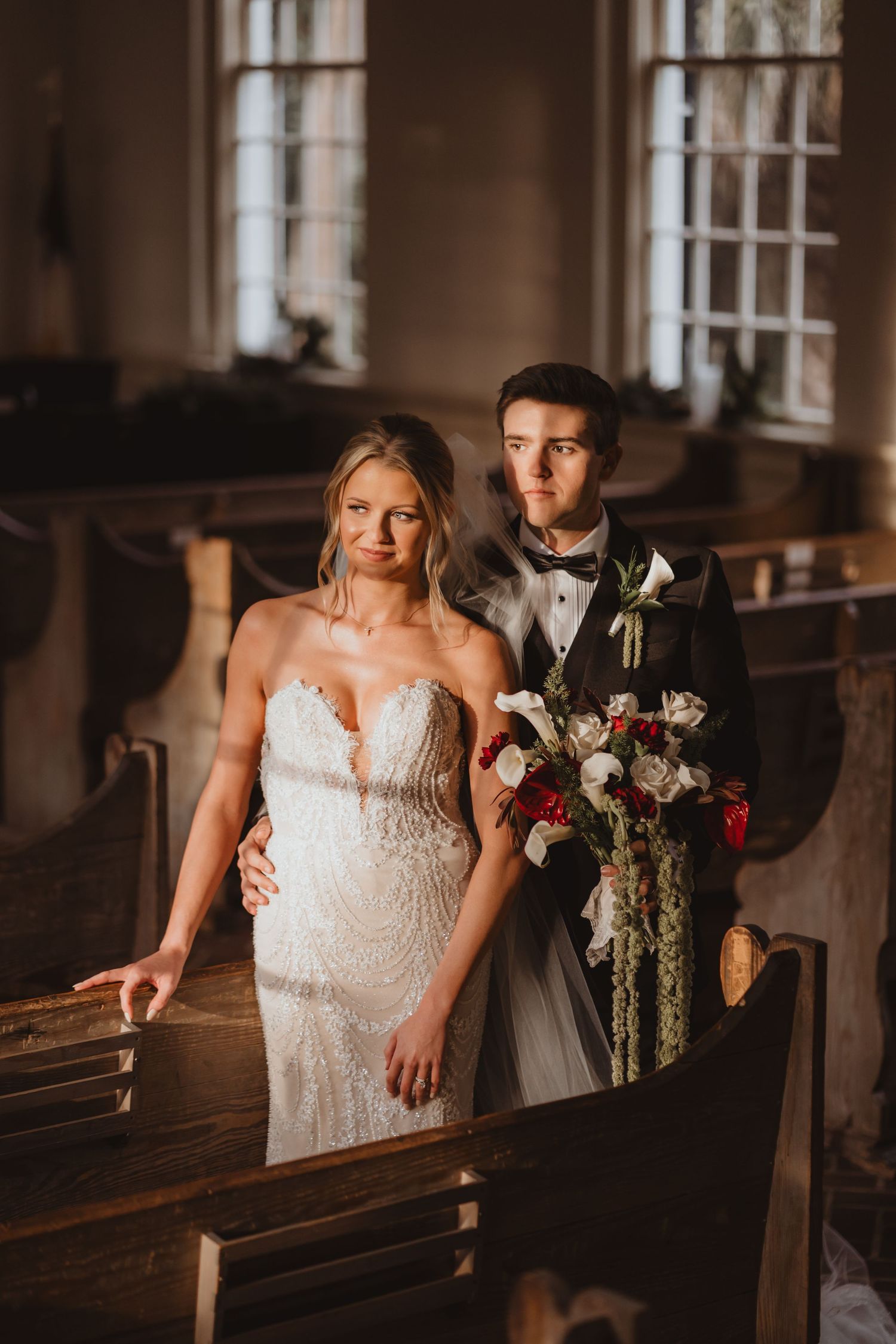 Bride in beaded strapless gown and groom in tuxedo pose in a rustic church holding red bouquet.