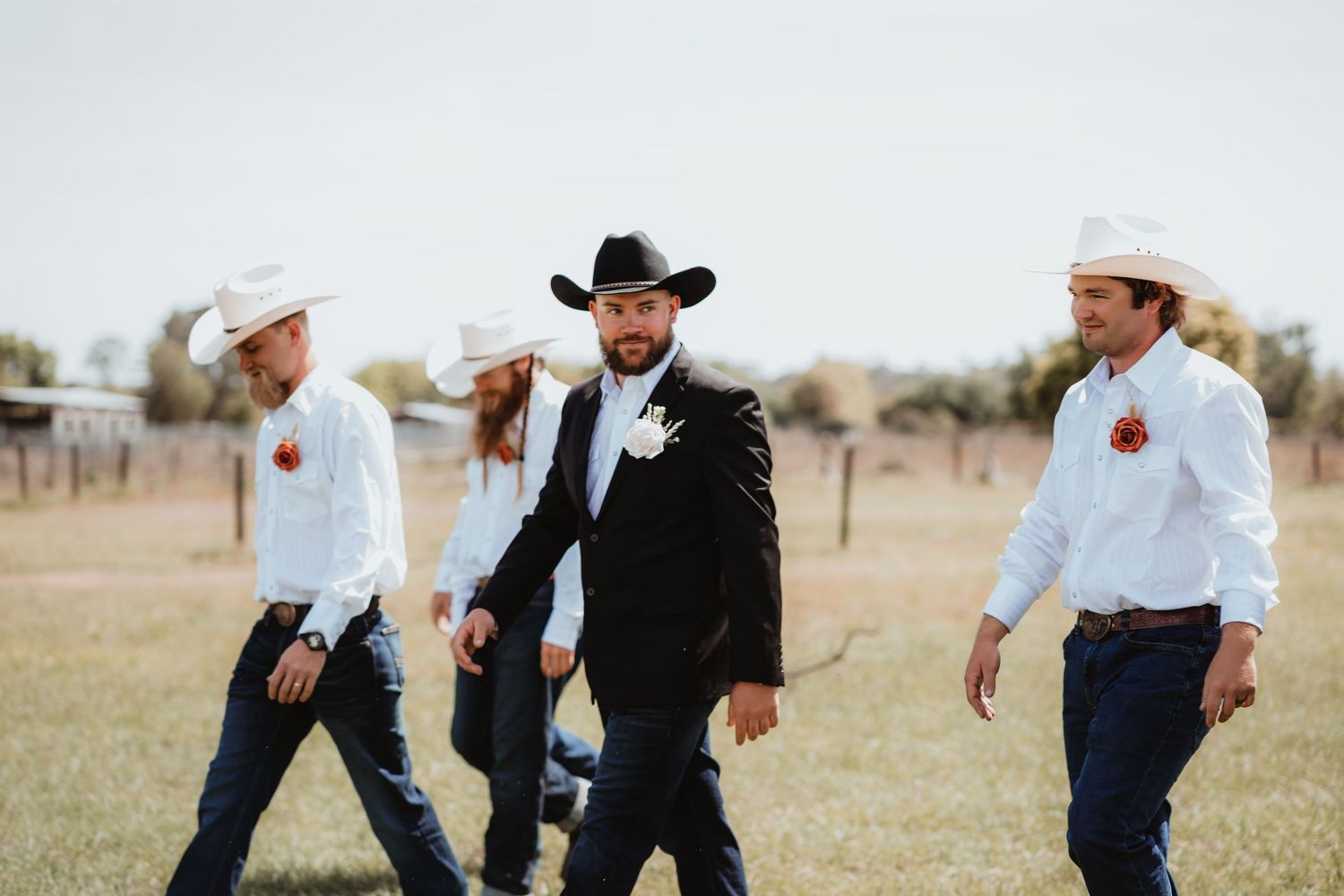 Groom wearing black cowboy hat and suit walks confidently with groomsmen at a country wedding venue.
