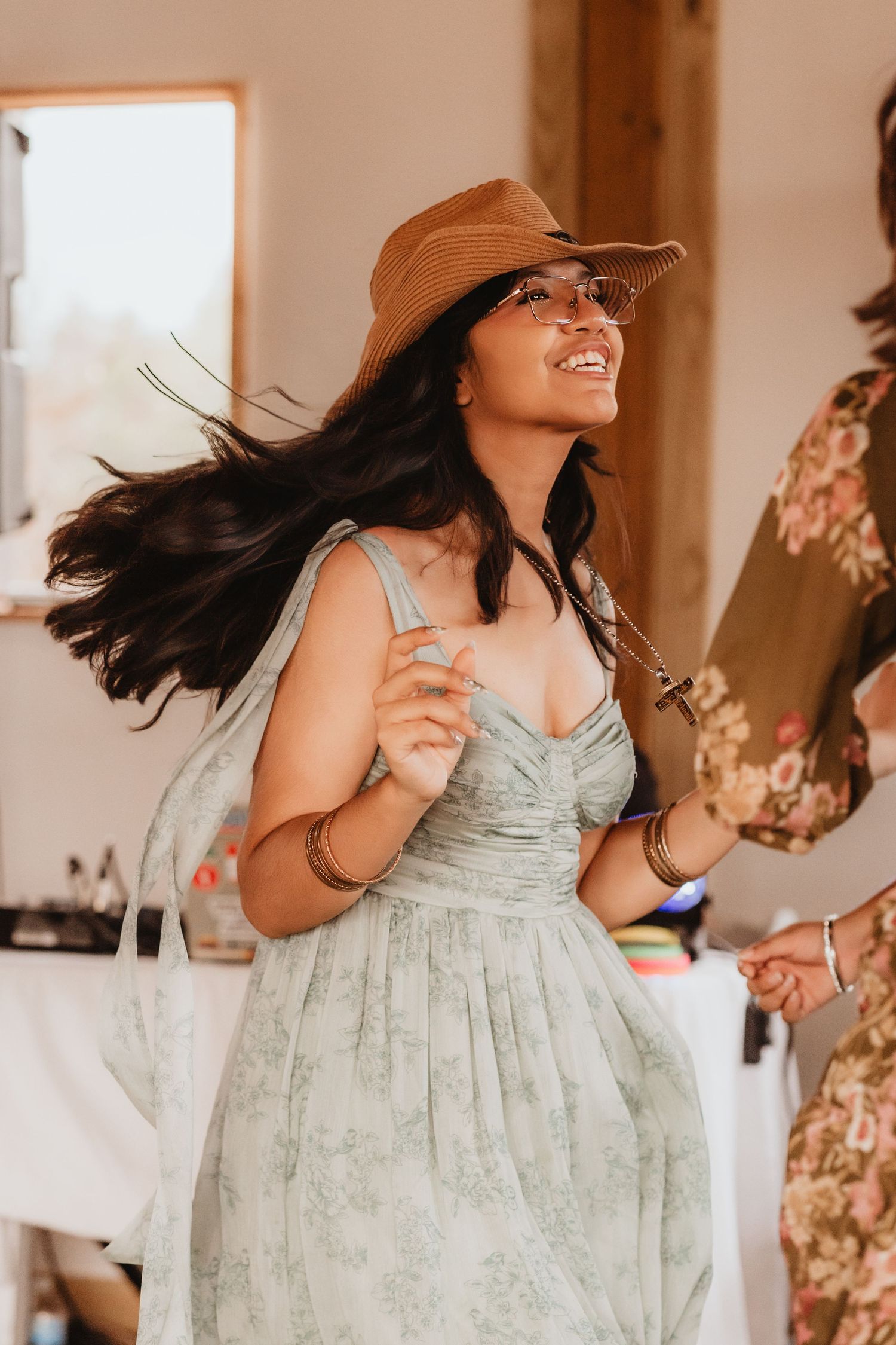 Woman in mint floral sundress and brown hat twirls joyfully at outdoor wedding reception.