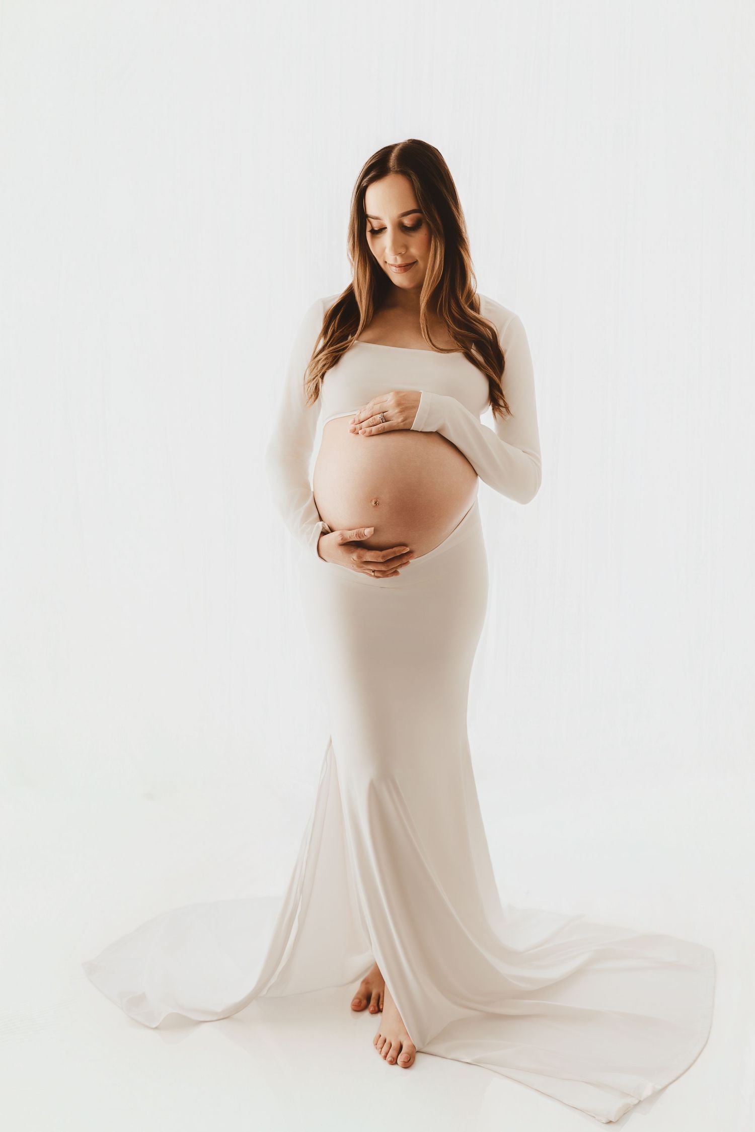 Elegant pregnant woman in a flowing white off-shoulder gown cradling her baby bump against a white background.
