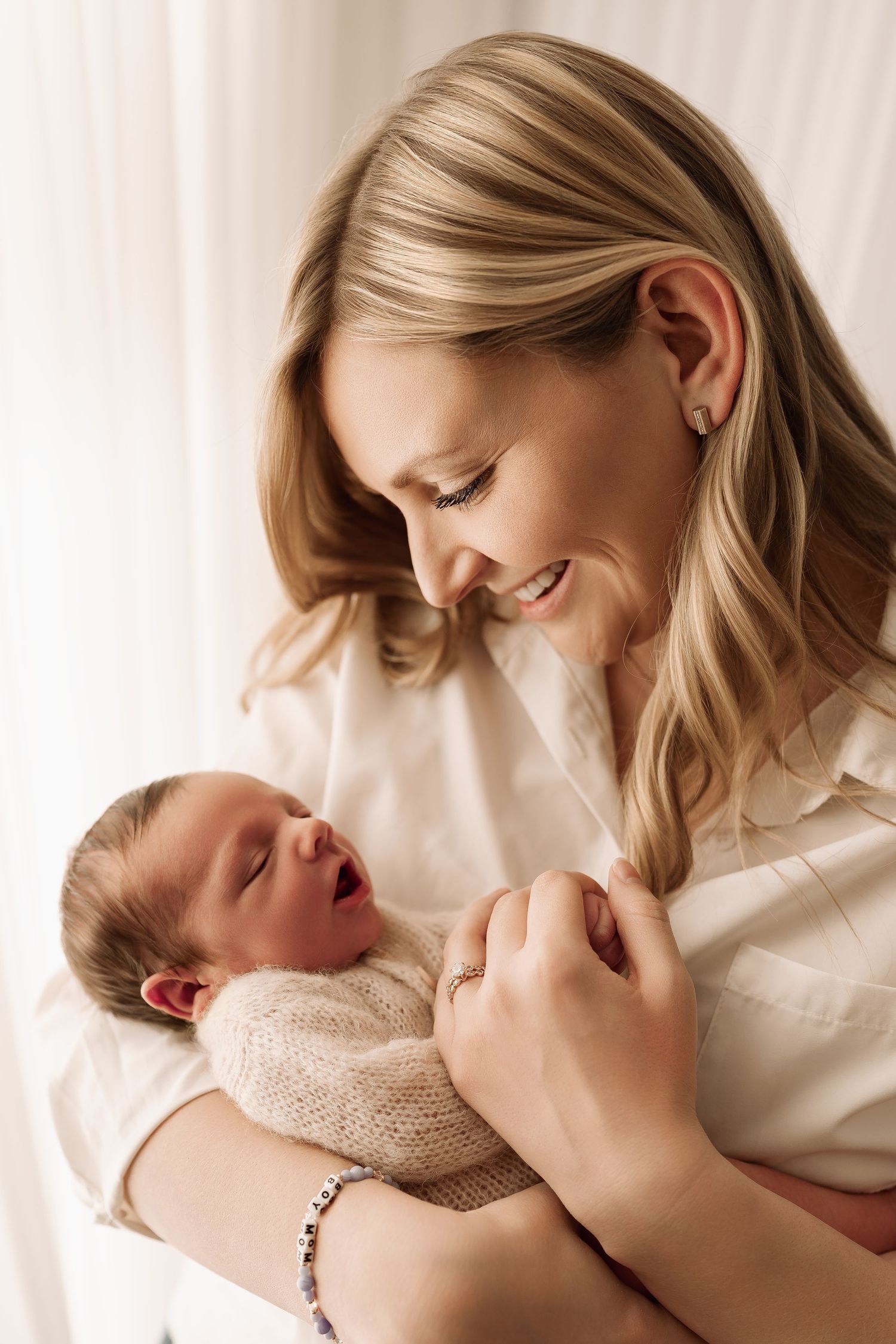 Blonde mother smiling warmly at her crying newborn baby wrapped in a knit blanket.