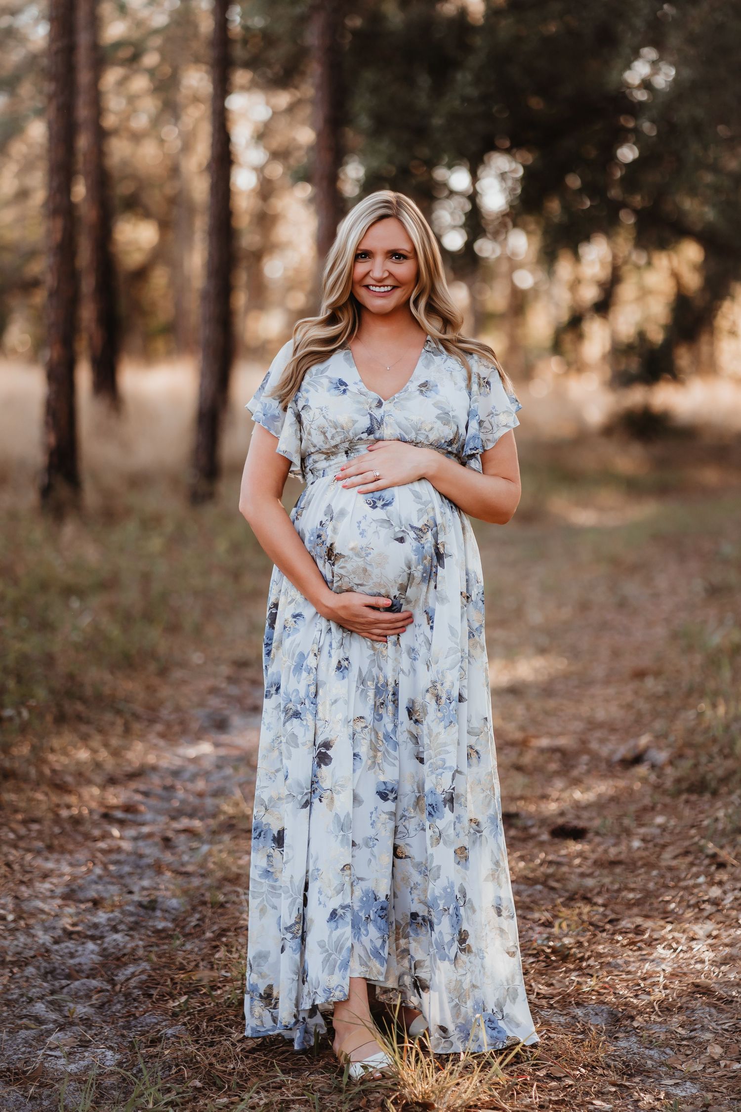Pregnant blonde woman in blue floral maxi dress smiling while holding baby bump outdoors.