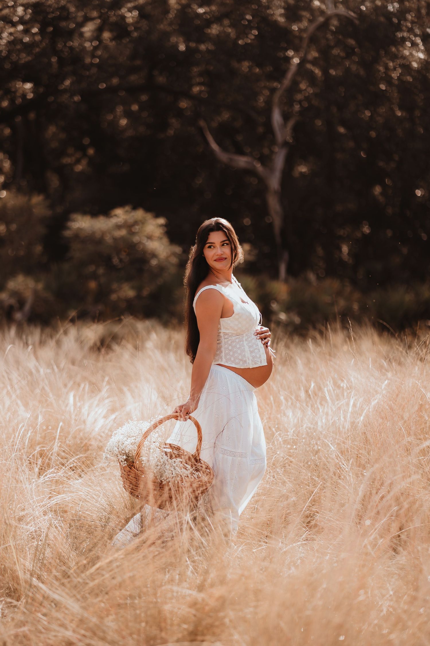 Smiling pregnant woman in white dress cradling baby bump while holding basket in golden meadow.