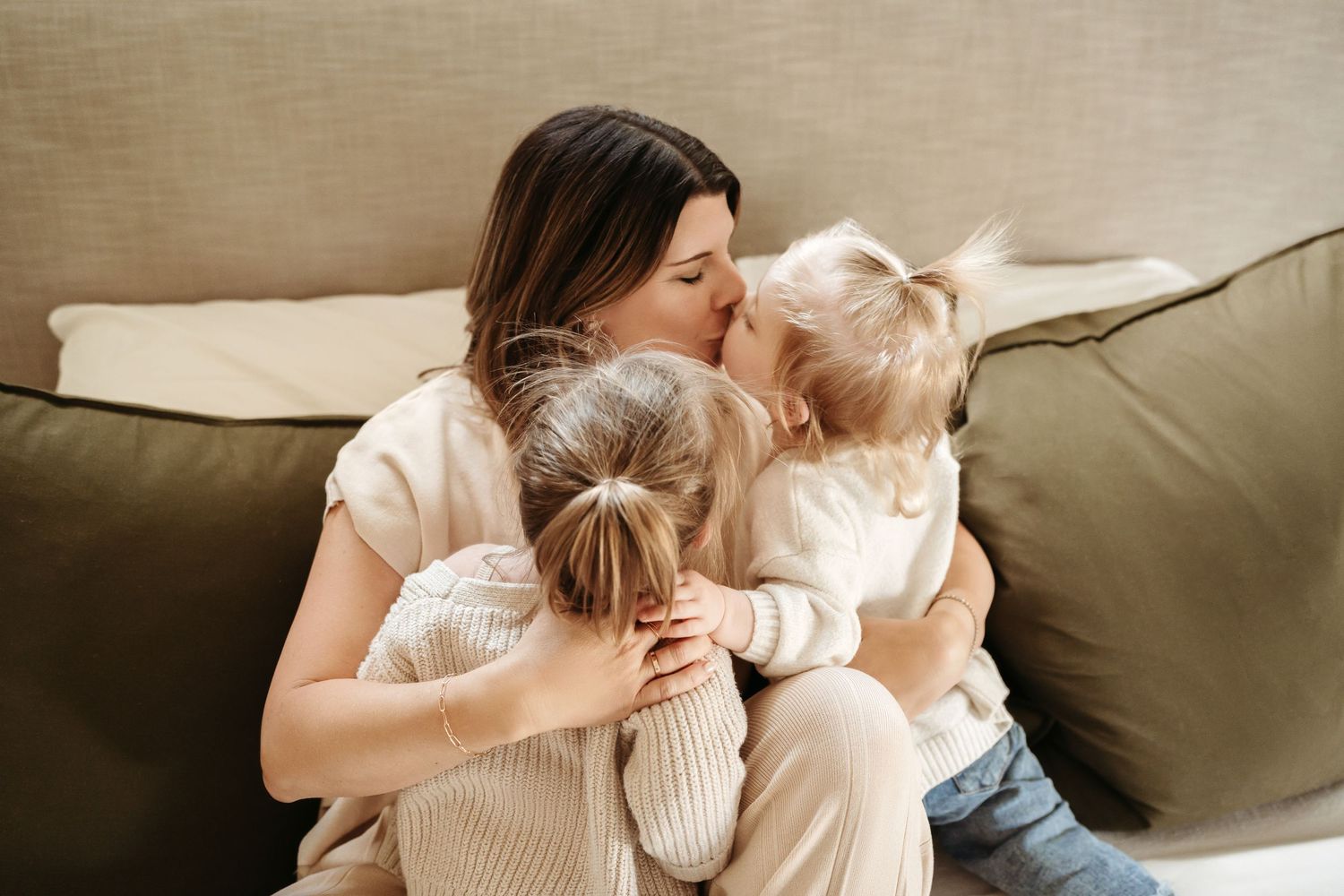 A dark-haired mother hugs and kisses two young blonde children on a cozy beige sofa.
