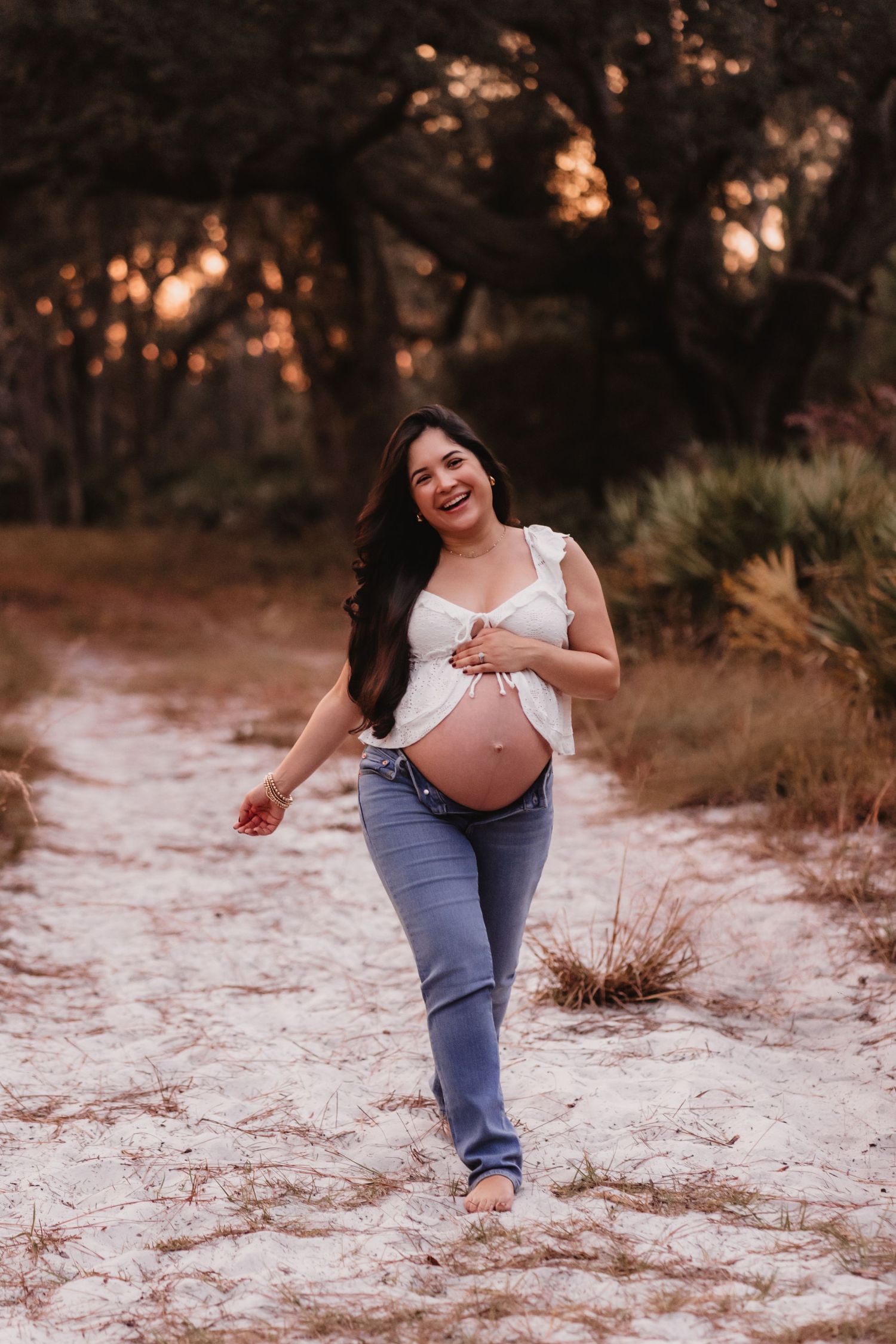 Smiling pregnant woman in white top and jeans walking barefoot on sandy path surrounded by trees at sunset.