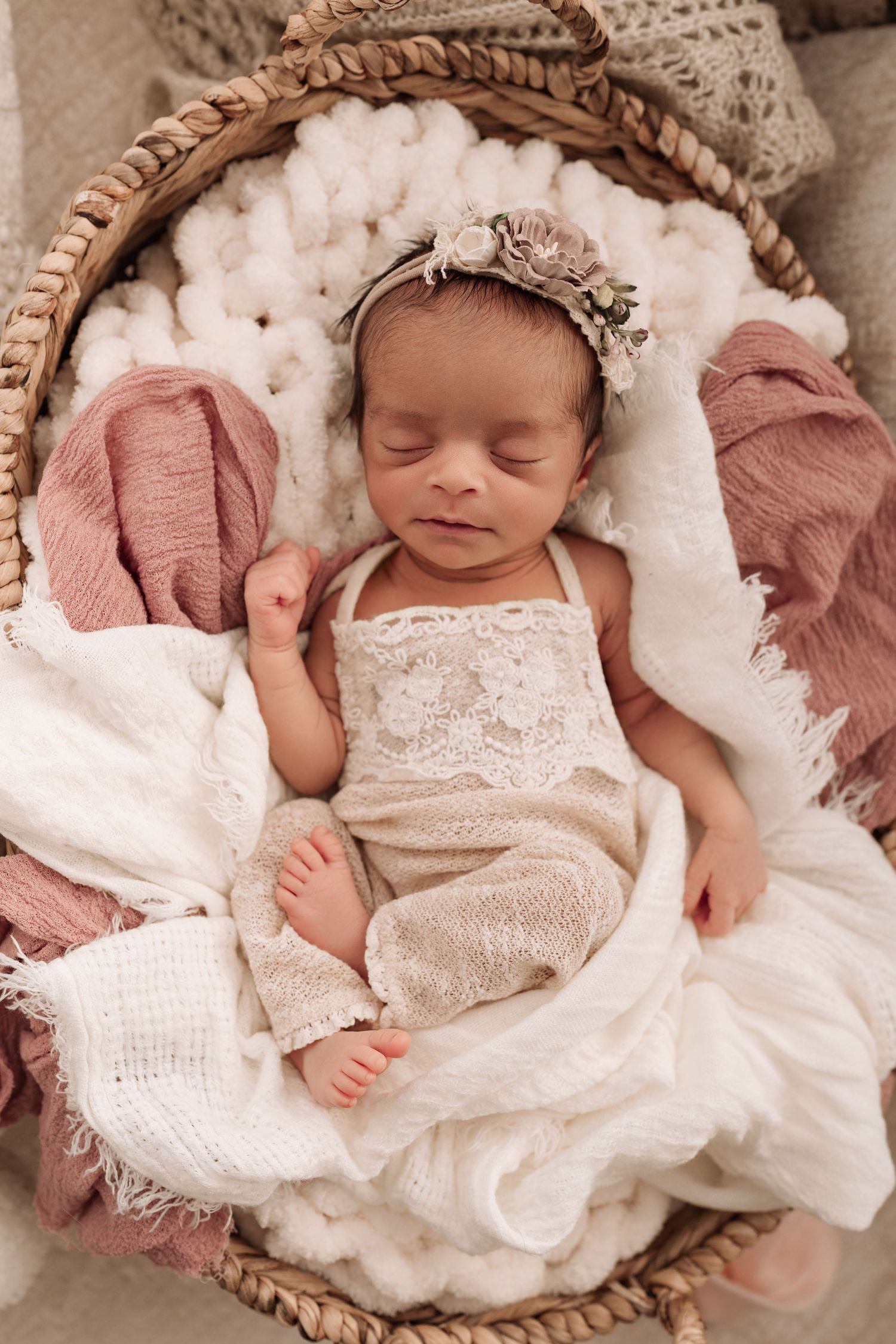 Newborn baby sleeping peacefully in a wicker basket with white and pink blankets, wearing a lace outfit and floral headband.